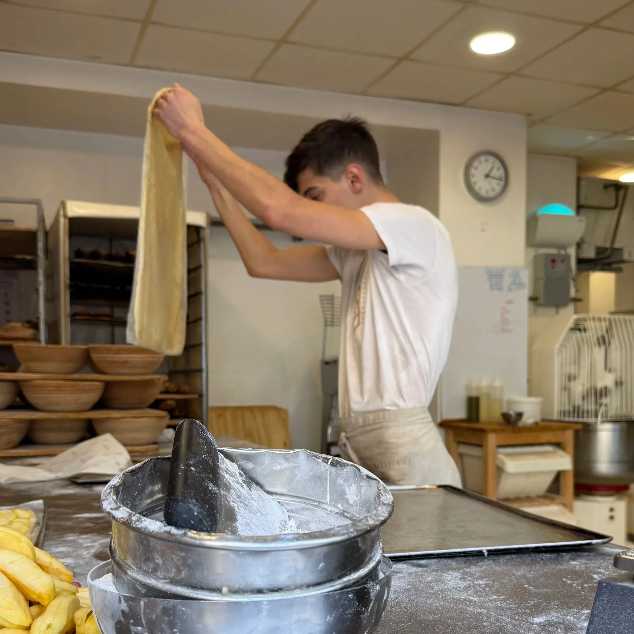 Un joven amasando eón el obrador de una panadería, sosteniendo y extendiendo una pieza de masa en el aire.