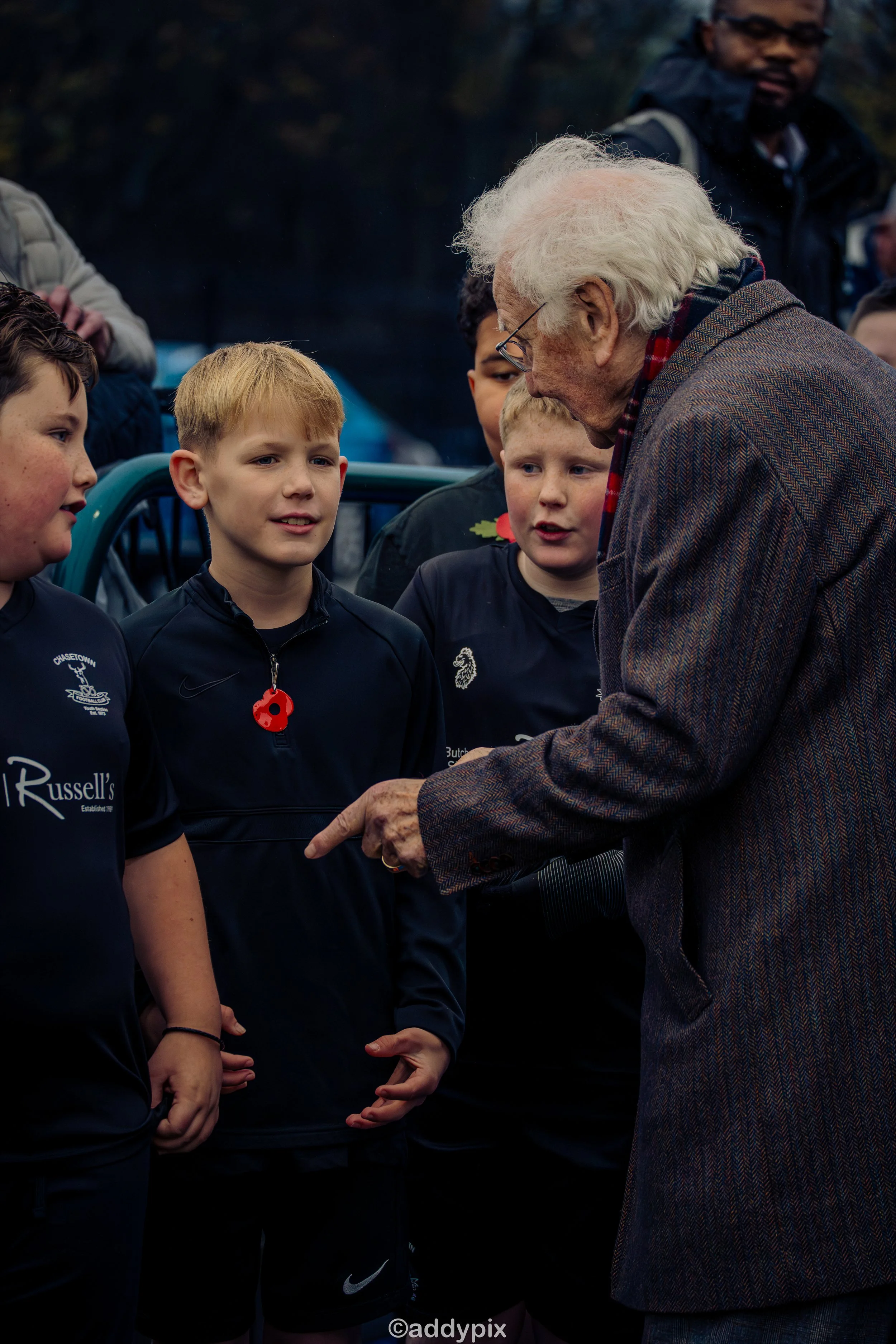 An elderly man with white hair talking to a group of young boys, in sports uniforms, outdoors during daytime.