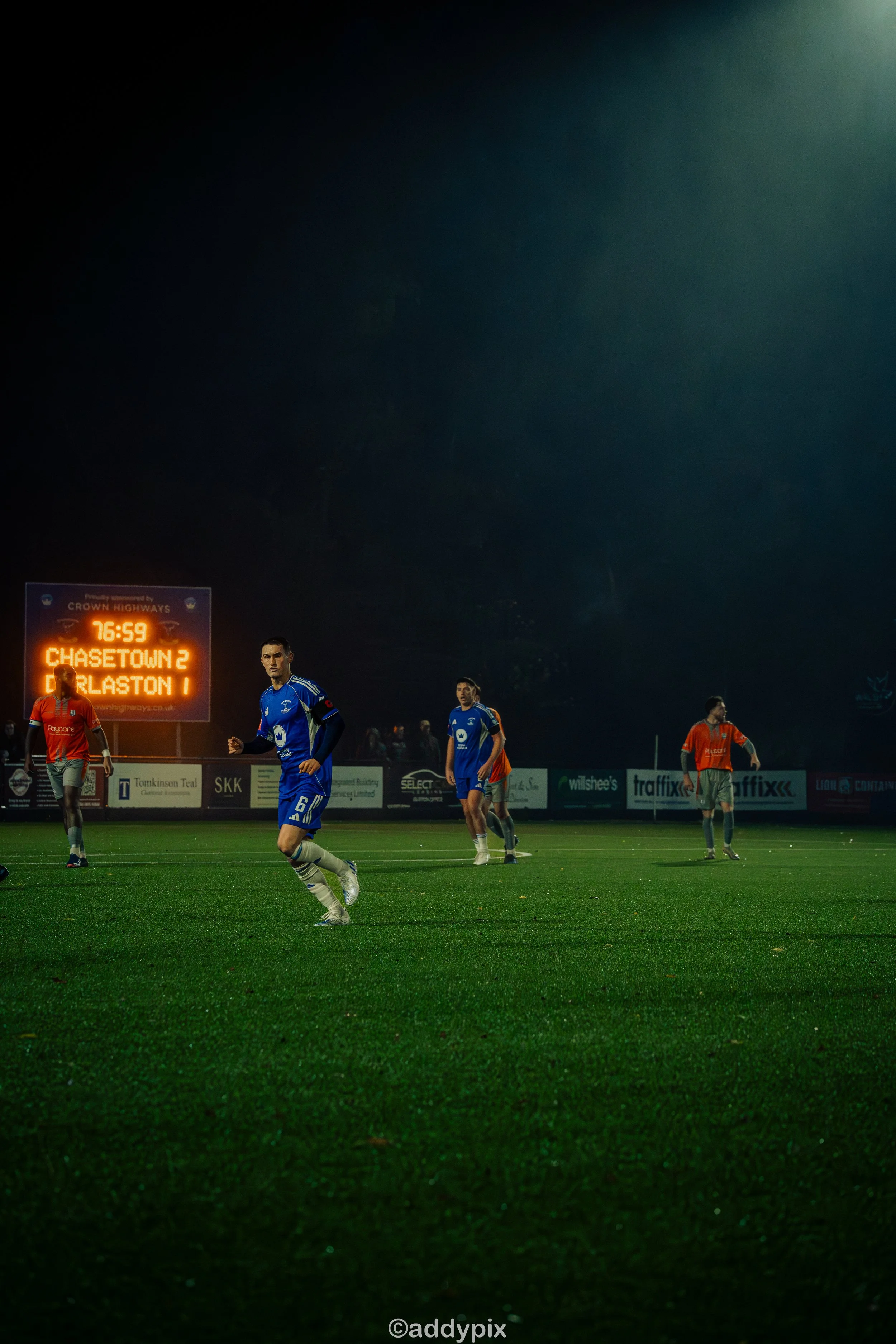 Soccer players on a field during a match at night with an electronic scoreboard in the background showing the score and time.