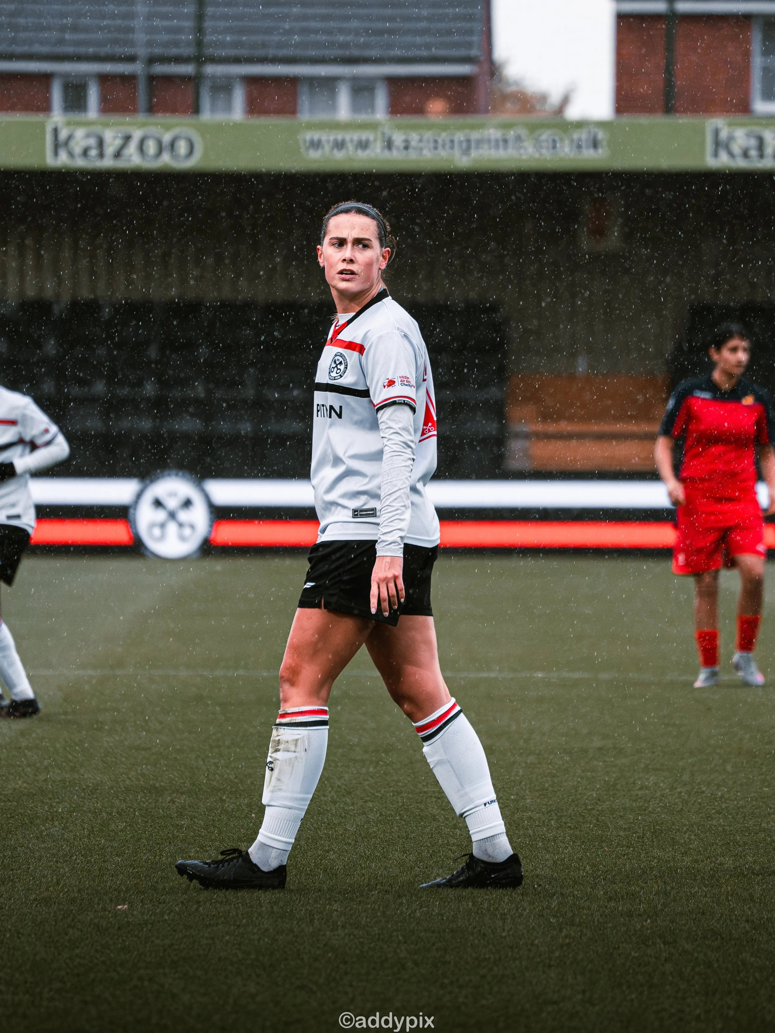 A female soccer player in a white and black uniform standing on a soccer field during a game in rainy weather. There are other players in the background, and a sign with the text 'kazoo' above.