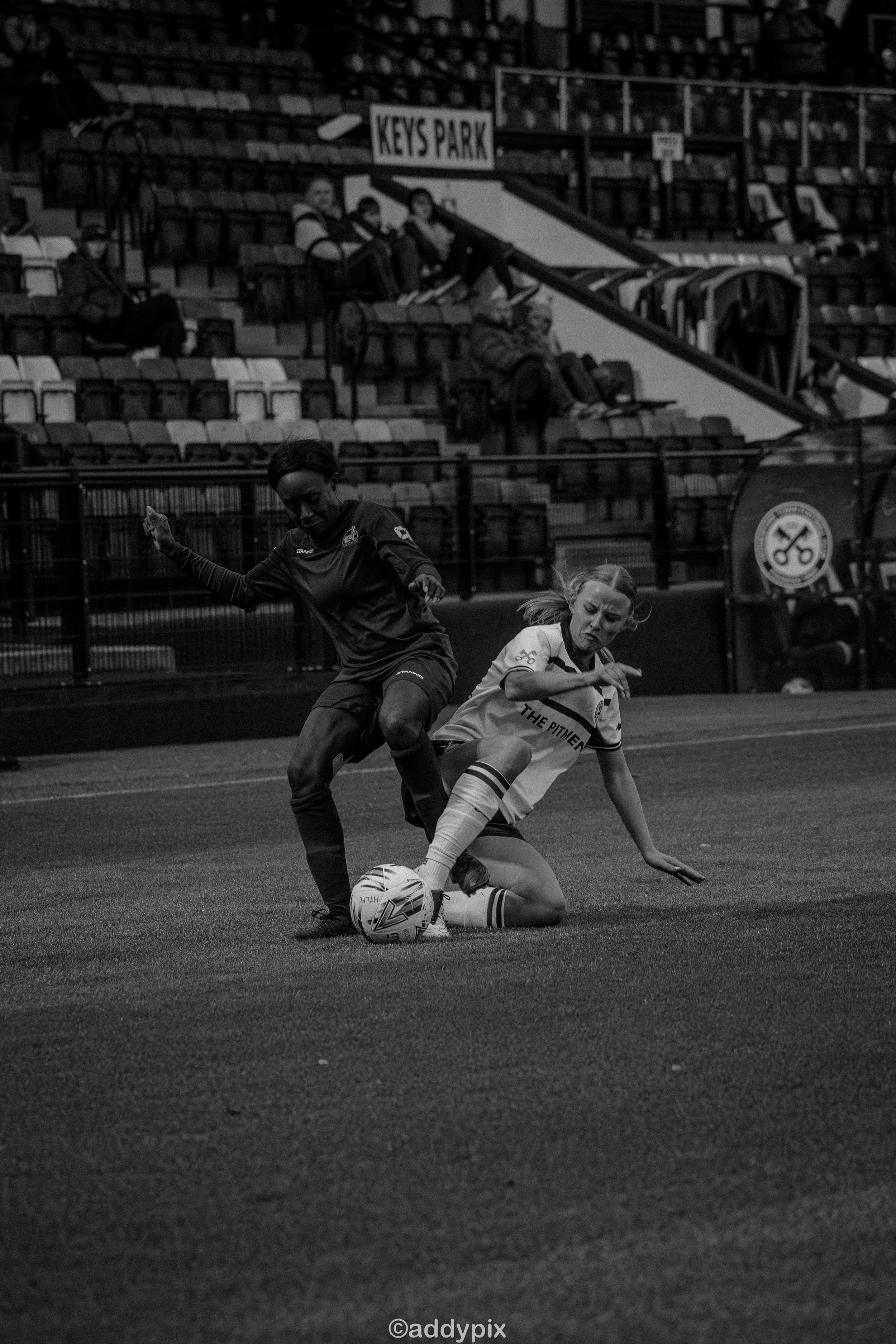 Two women playing soccer on a field, one is on the ground and the other is standing, contesting for the ball, with empty stadium seats in the background.