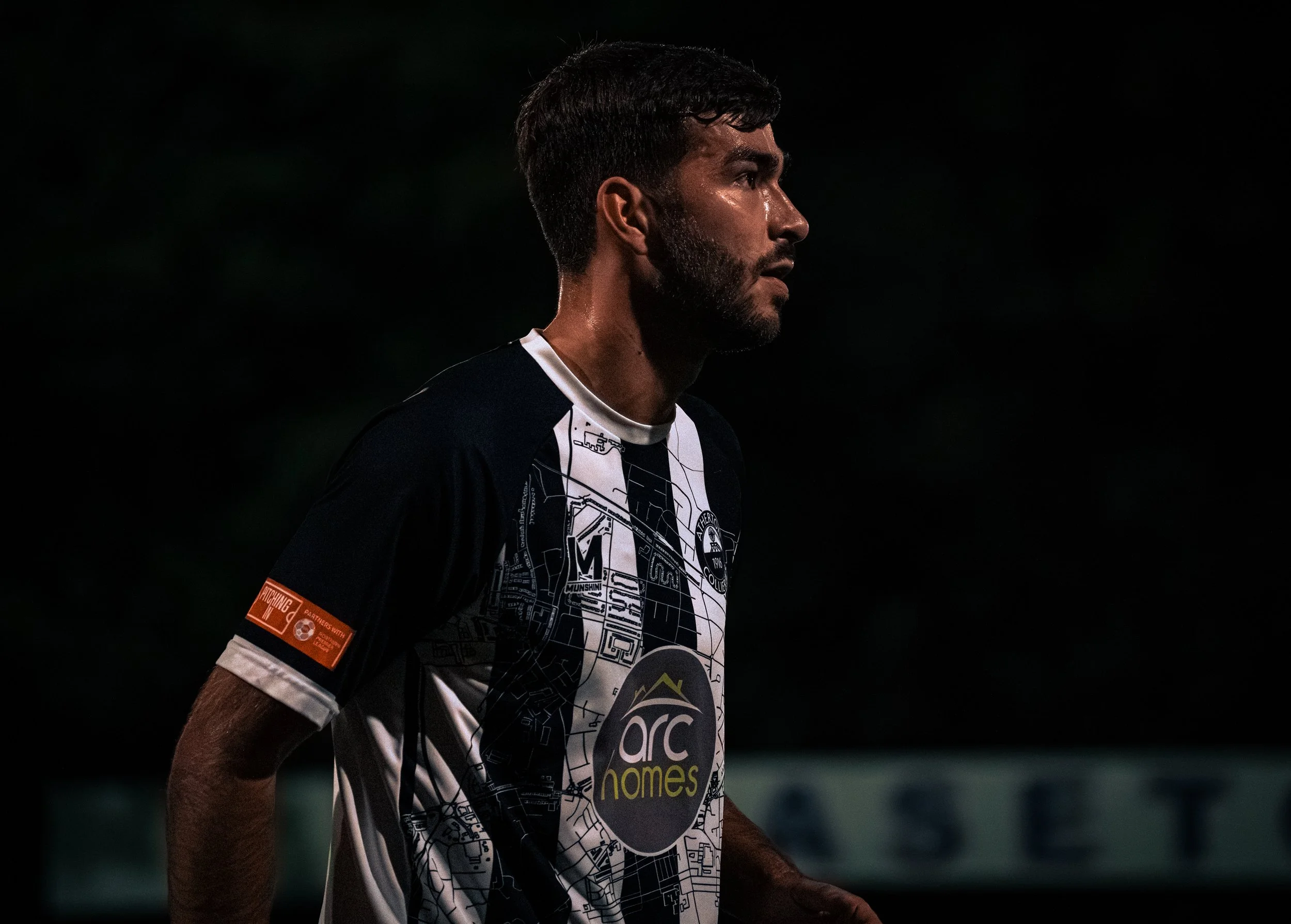 A male soccer player with dark hair and a beard standing on the field at night, wearing a black and white jersey with a map design and sponsor logos.