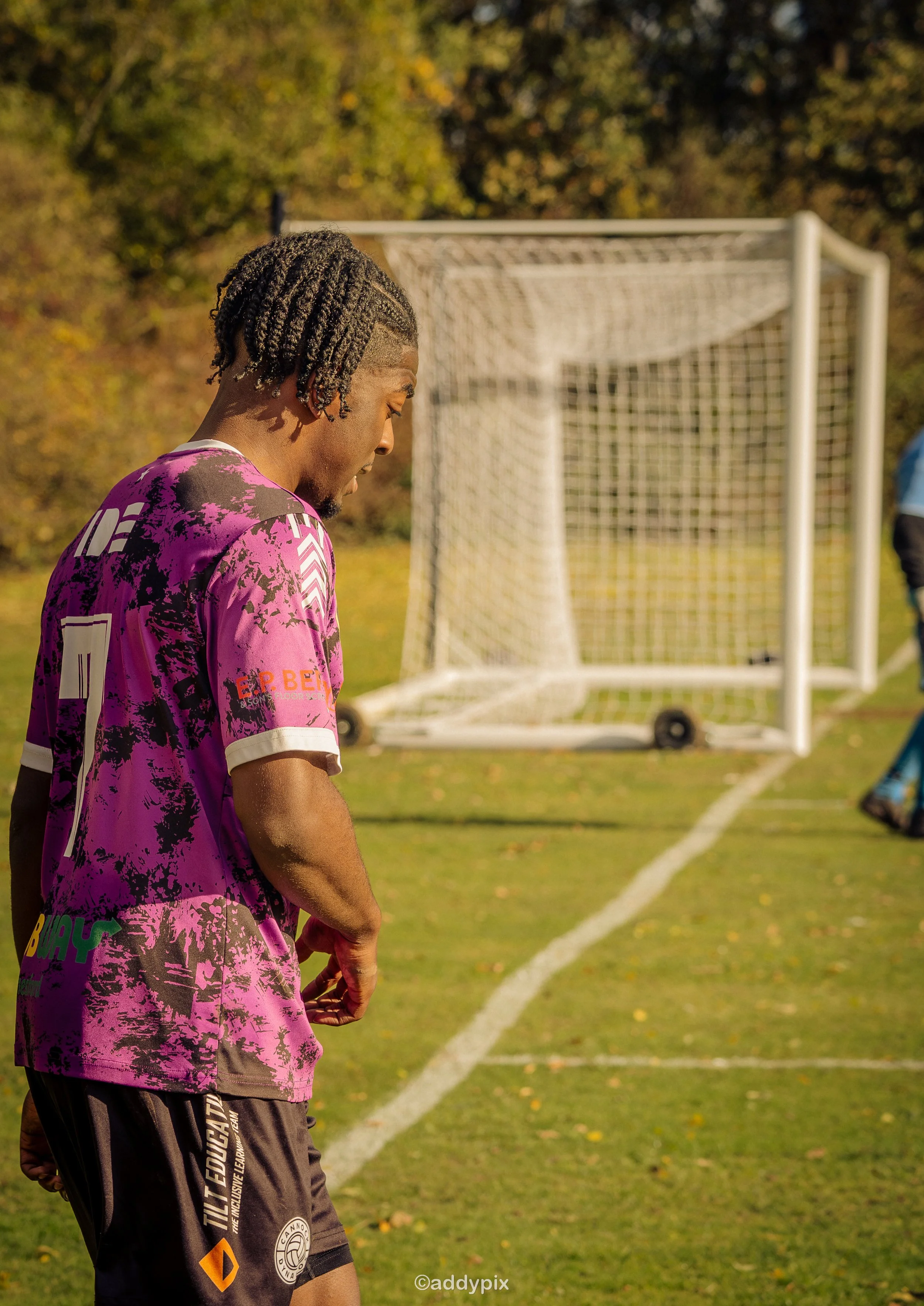 A soccer player in a pink and black jersey standing on the field with a soccer goal in the background during daytime.
