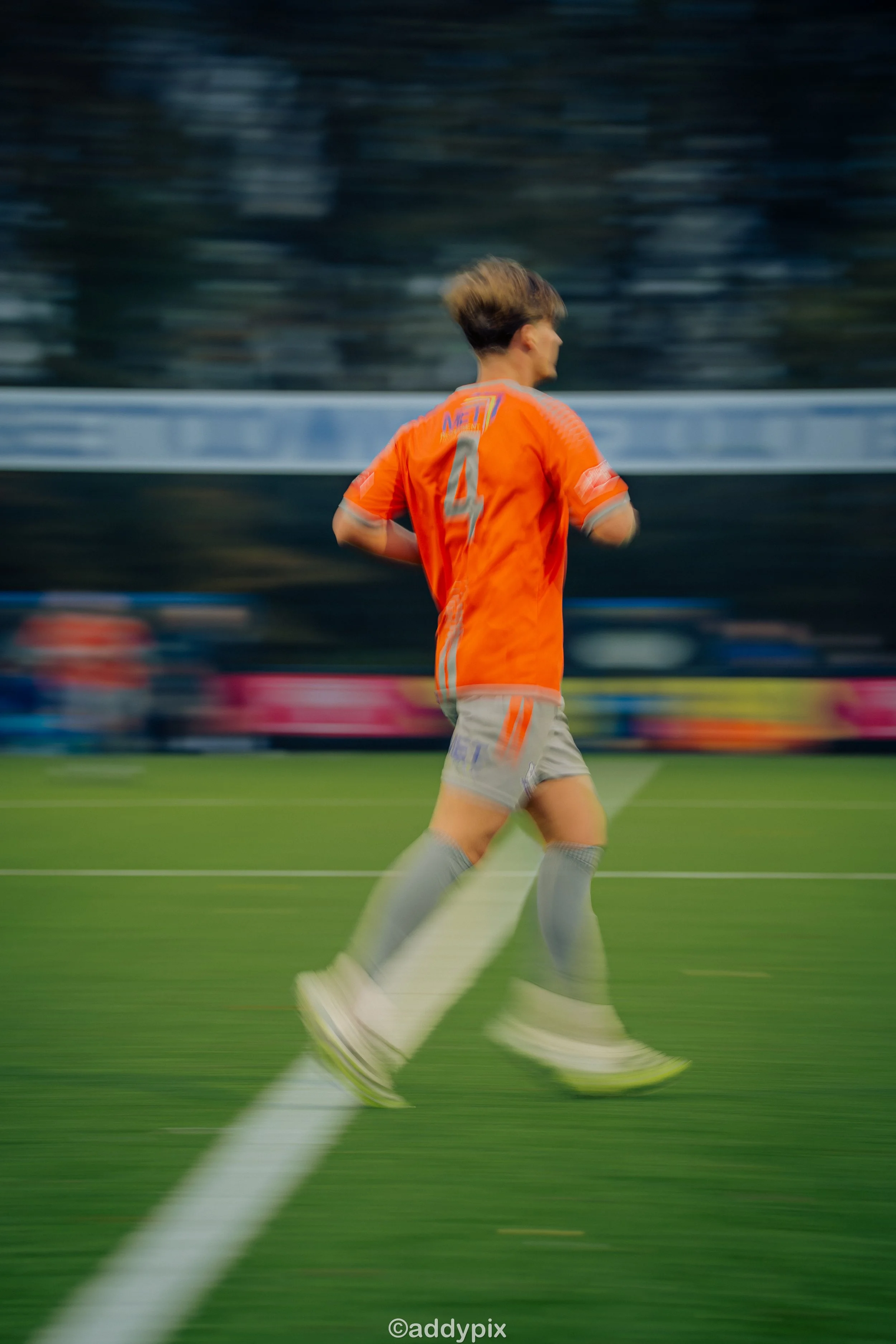 A soccer player wearing an orange jersey with the number 4 and gray shorts, running on a green field during a game, with motion blur.