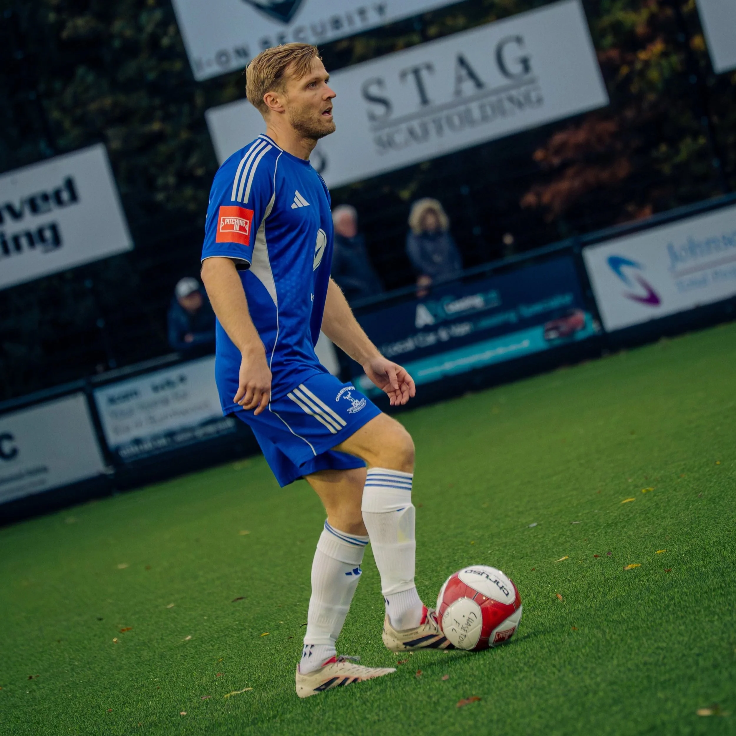 A soccer player in a blue uniform preparing to kick a red and white soccer ball on a field, with advertising boards and people in the background.