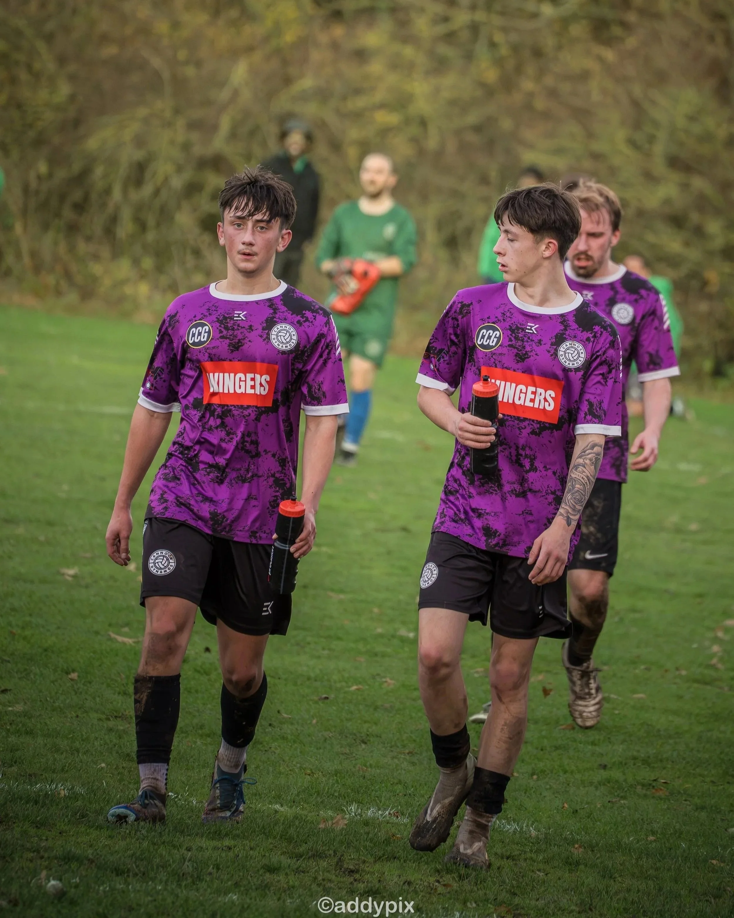 Young male soccer players in purple jerseys walking on the field with water bottles, with others in the background.