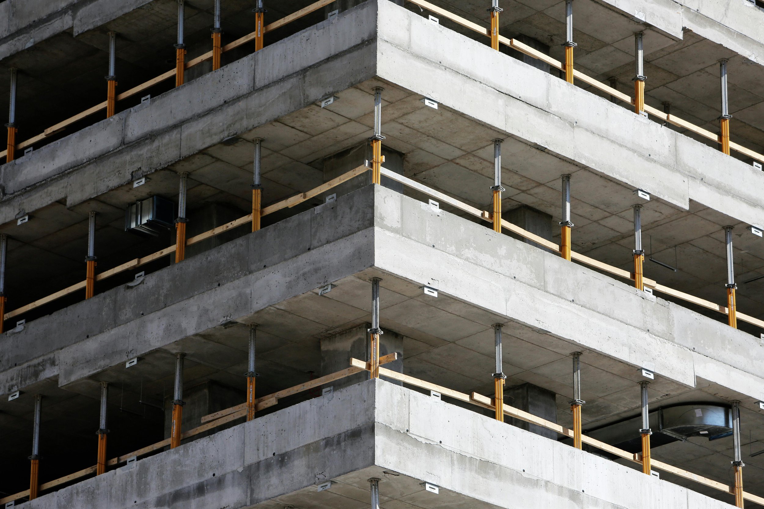 Several floors of a concrete building under construction, with exposed concrete slabs and orange scaffolding supports.
