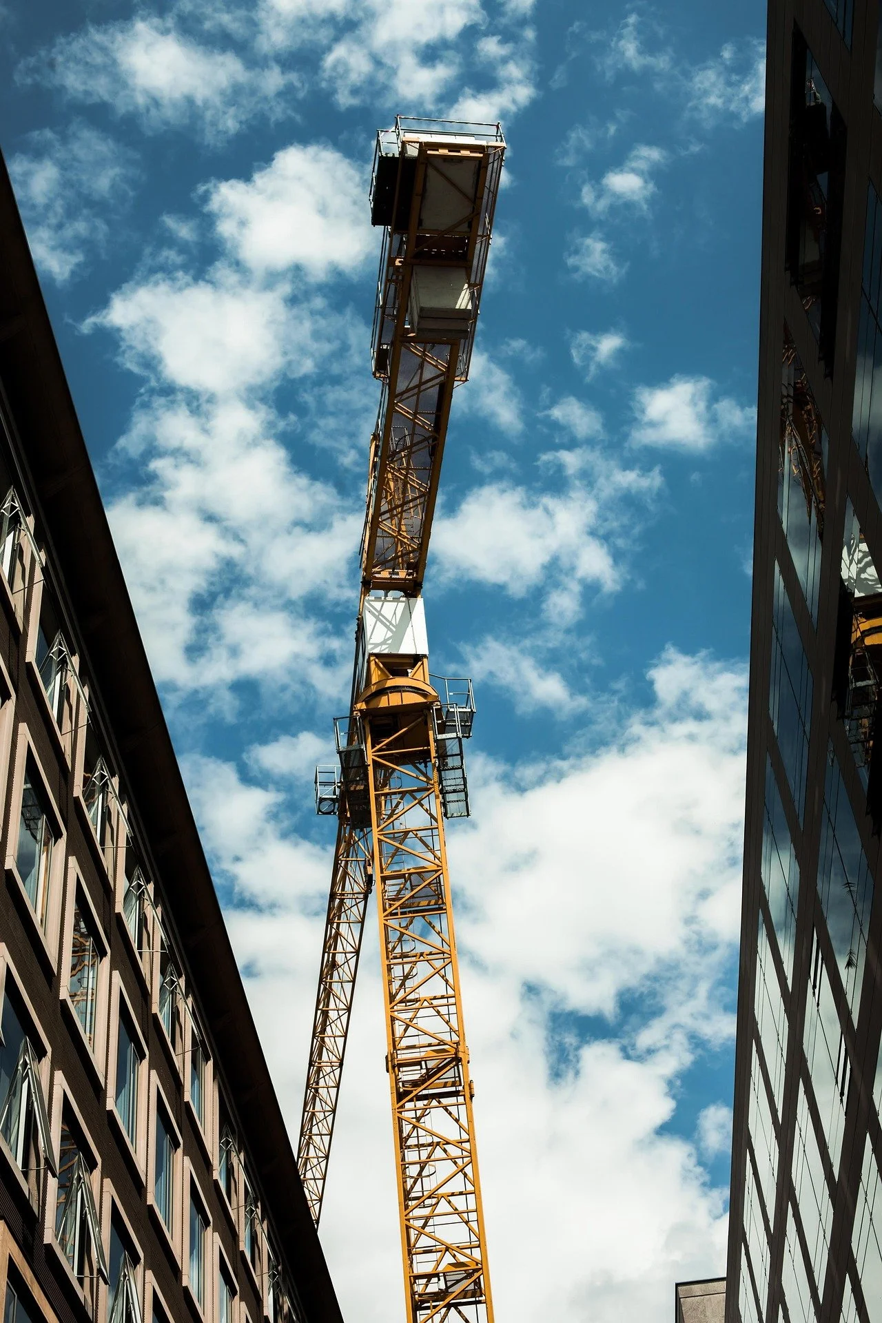 A yellow construction crane extending upward between two modern buildings with reflective glass windows, against a blue sky with scattered white clouds.