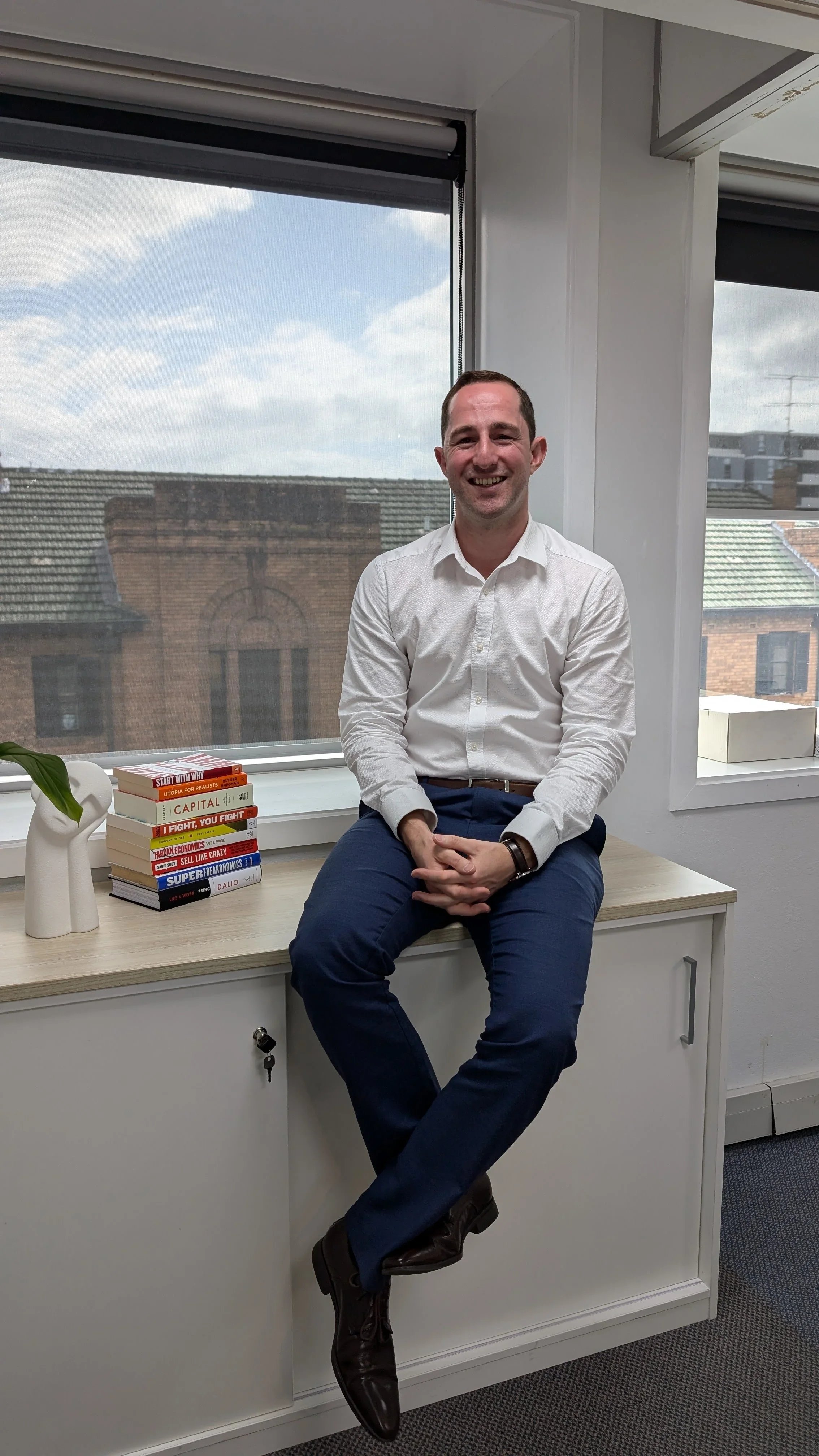 Leland from TradeScale Accountants and Advisors in a white shirt and dark pants sitting on a desk in an office, smiling, with books on the desk and windows showing rooftops and a cloudy sky in the background.