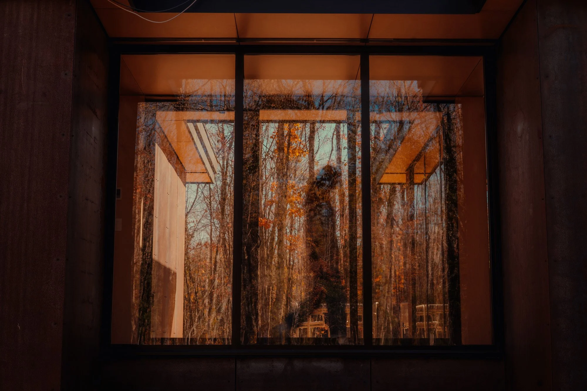 View through a large glass window, reflecting trees with autumn leaves and a person standing outside.