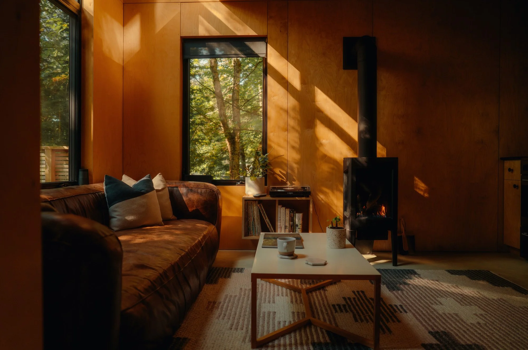 A cozy living room with wooden walls, a leather sofa with cushions, a white coffee table with a cup and remote on it, a small bookshelf with books, a potted plant, and a wood-burning stove with a fire. Sunlight streams through large windows showing t