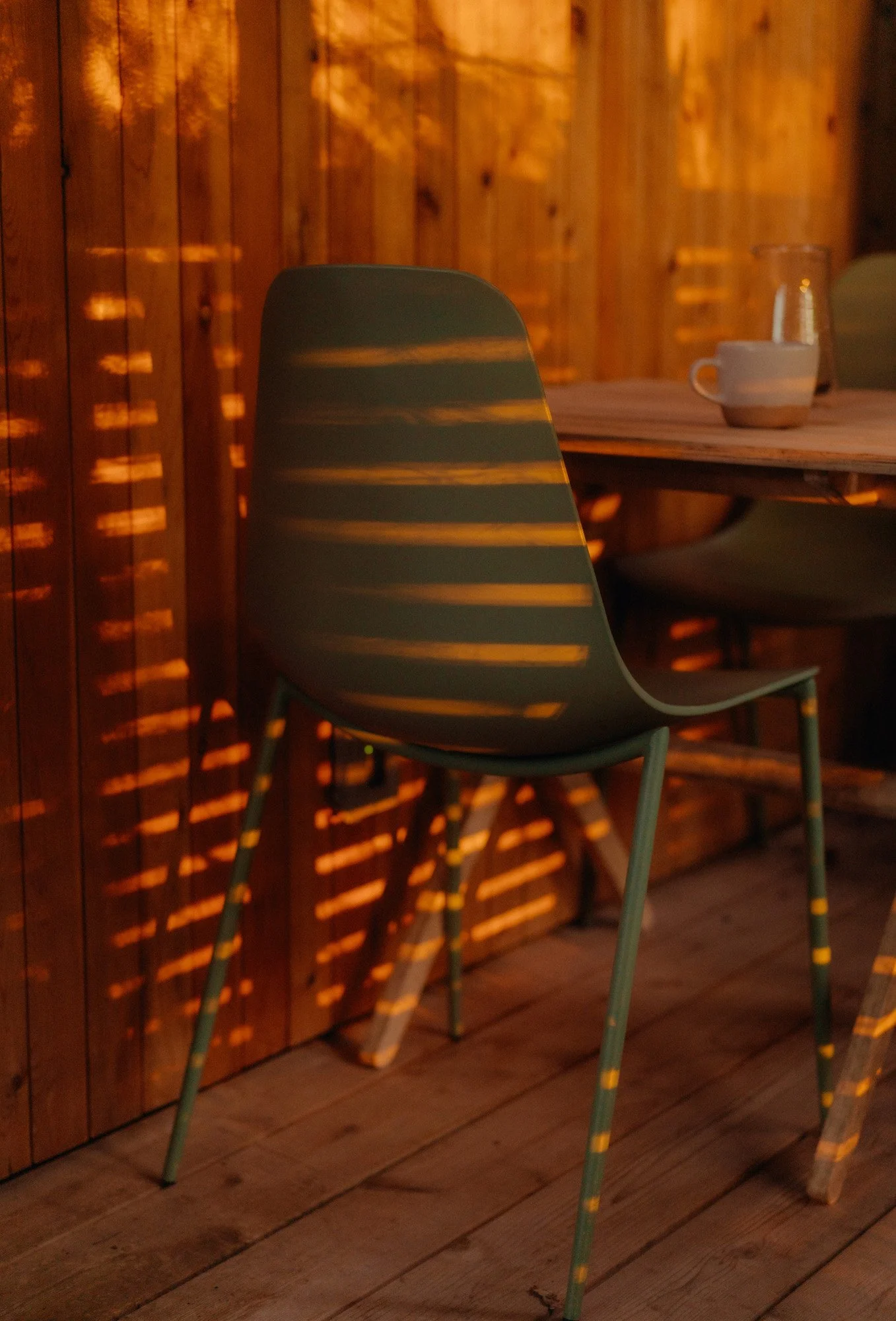 A green chair with light shining through a wooden slat wall, creating pattern shadows on the chair and the wooden floor. A table with a cup and a glass is partially visible in the background.