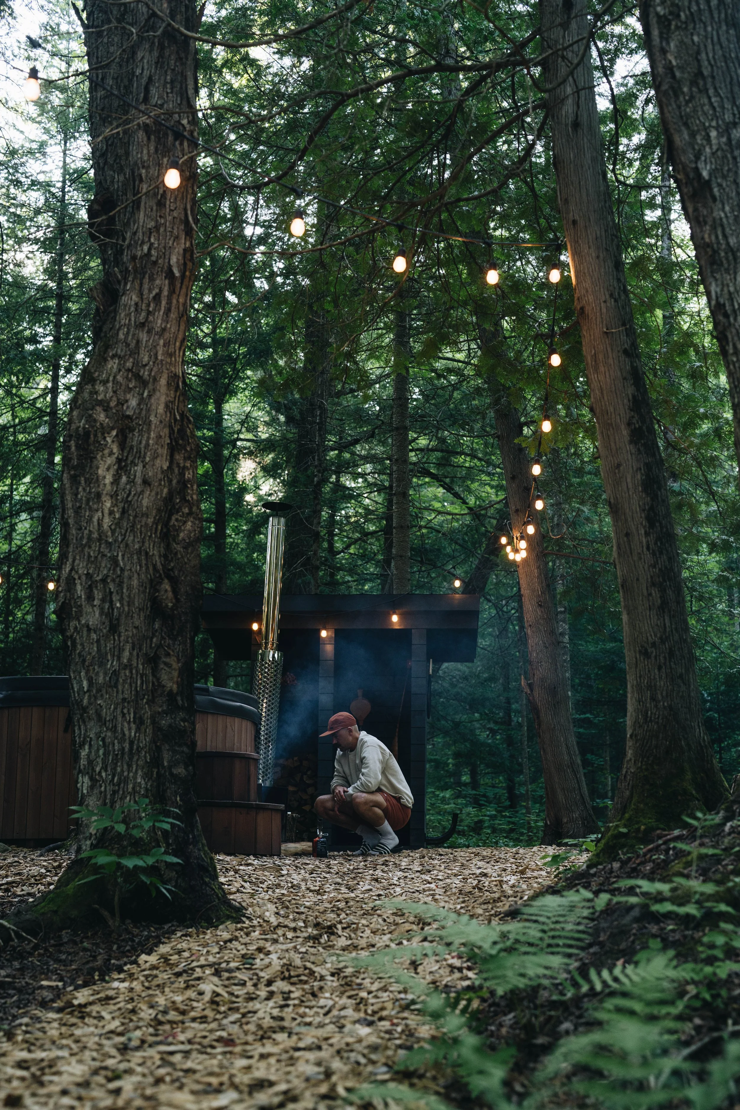 A person squatting in a forest clearing with a hot tub nearby and string lights hanging above.