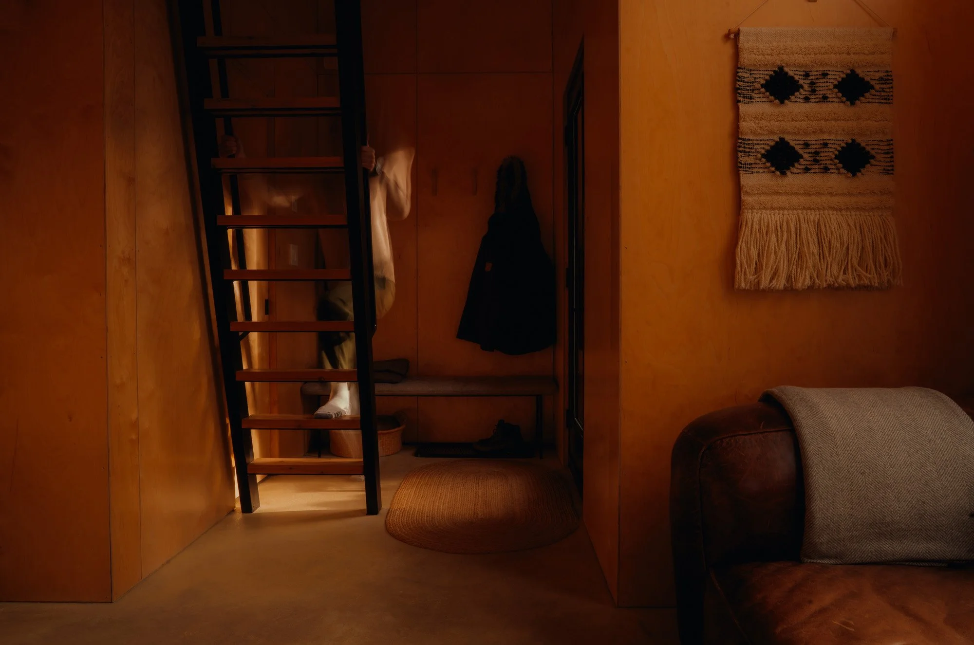 Interior of a cozy living space with warm lighting, a brown leather sofa with a gray cushion, a wall hanging with black designs and fringe, a small round rug, and a wooden staircase leading up to a loft or second level. There is also a coat hanging o