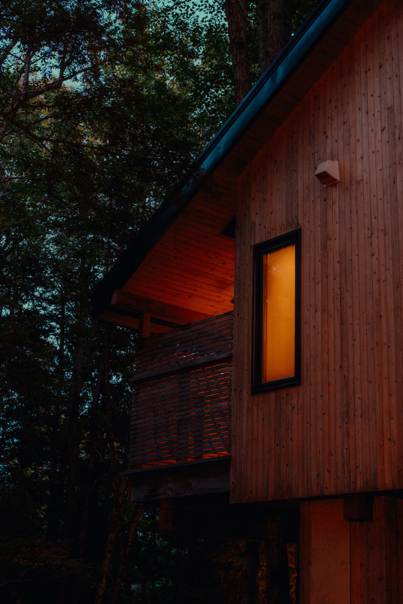 Nighttime view of a wooden house with warm light glowing from a window, surrounded by trees.