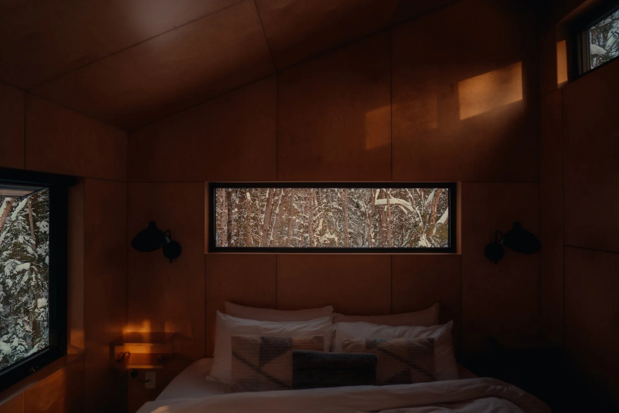 Interior of a bedroom with wooden walls and a bed, featuring two black wall-mounted lamps and a nightstand, with snowy trees visible through the horizontal window and side windows.