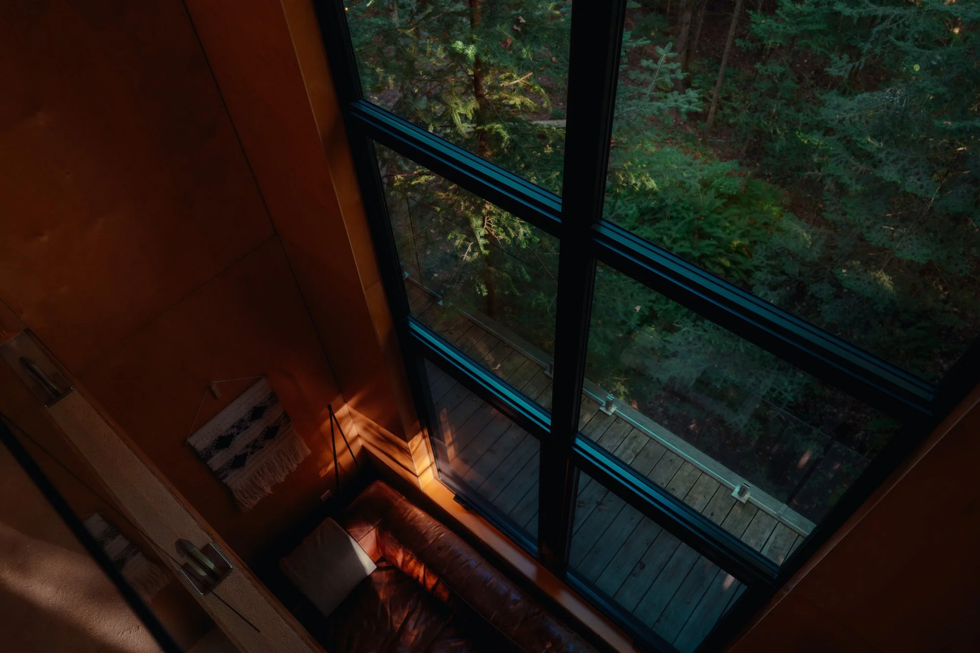 Interior of a modern room with a large glass window showing a view of lush green trees outside. Part of a leather sofa with a pillow and a woven wall hanging are visible.