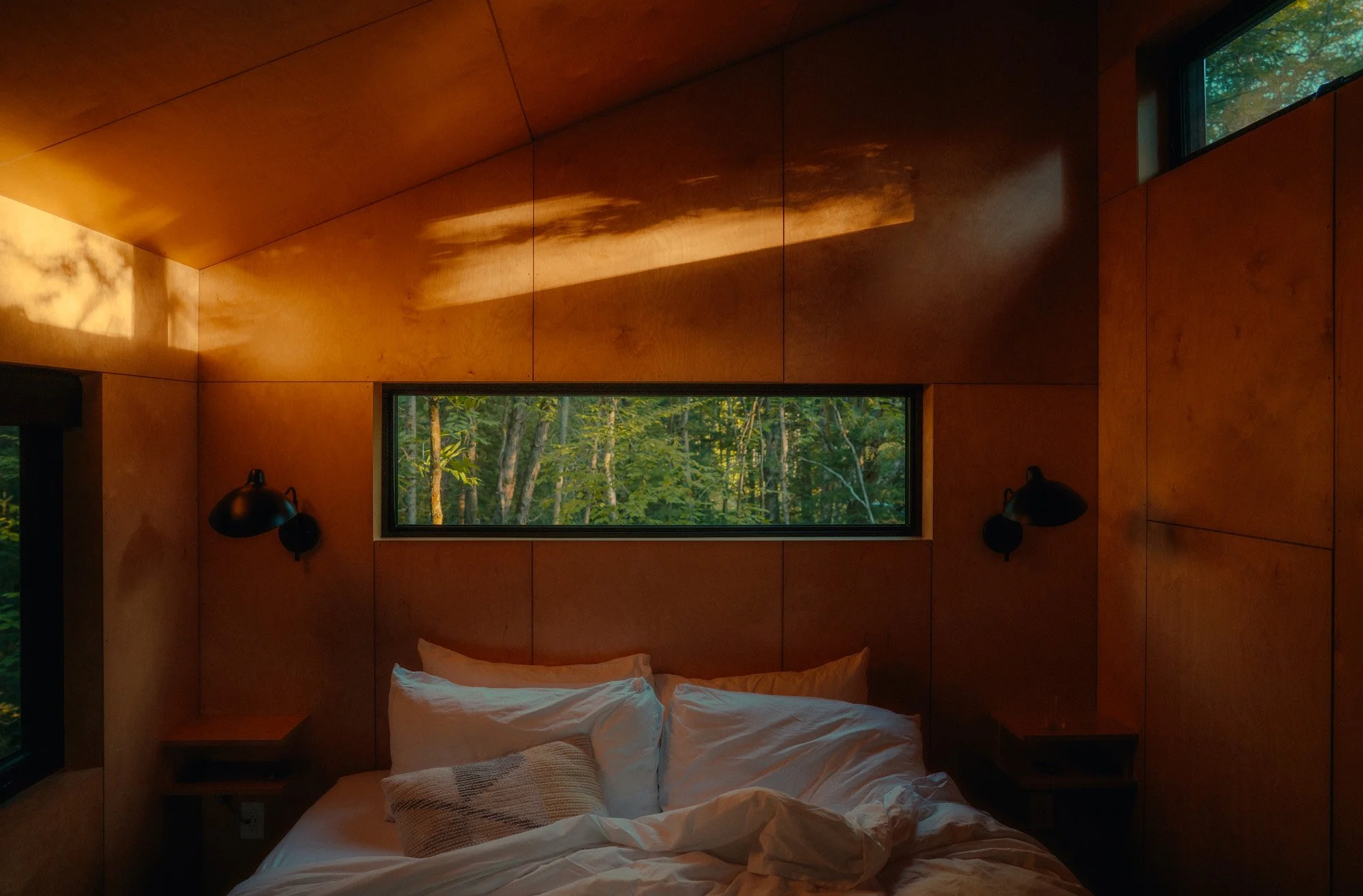 Cozy bedroom with wooden walls, a bed with white bedding and multiple pillows, and a horizontal window showing a view of trees outside.