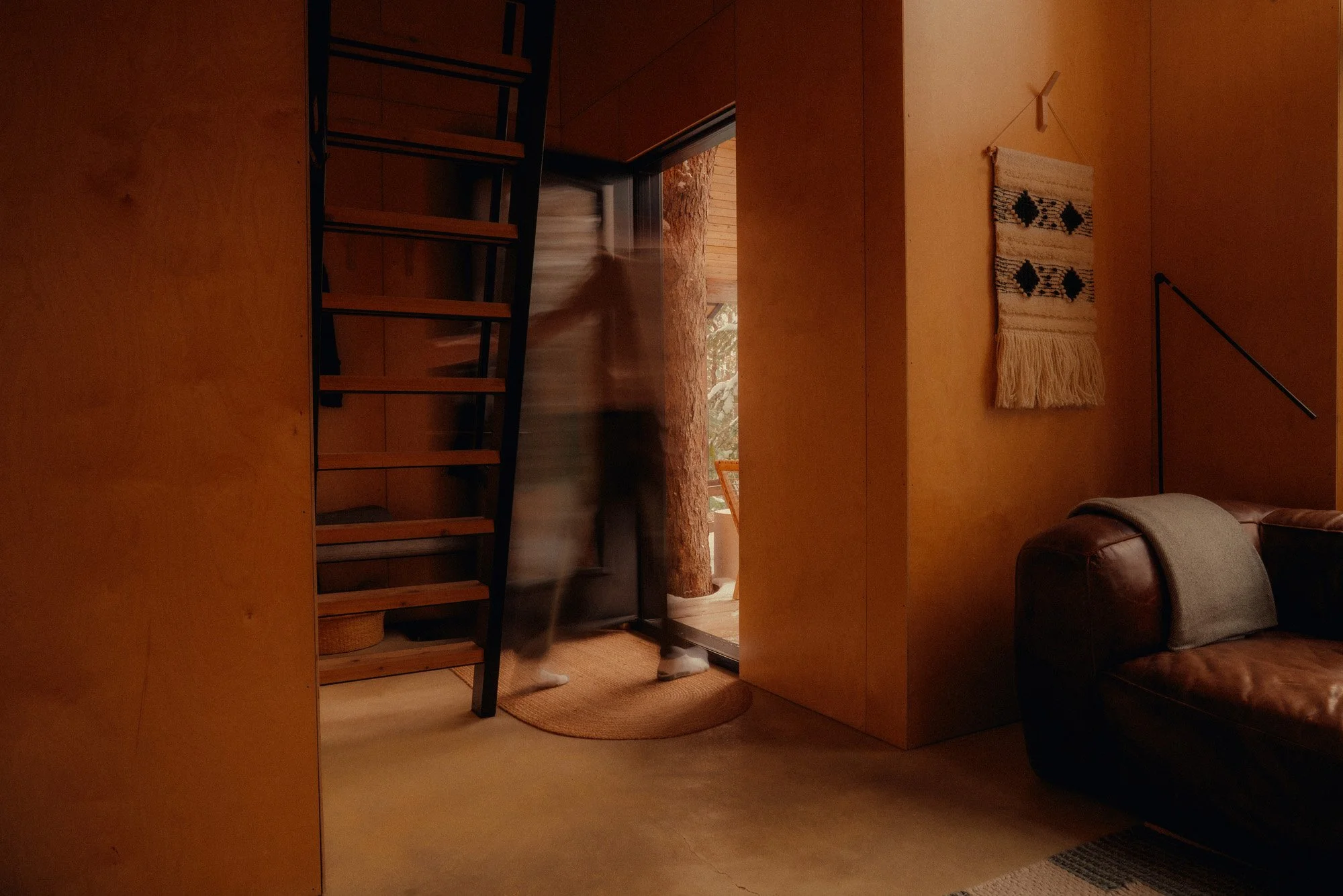 A living room with wooden walls, a leather couch with a blanket, a hanging textile wall decoration, a black floor lamp, and a staircase leading onto a balcony with a person stepping outside.