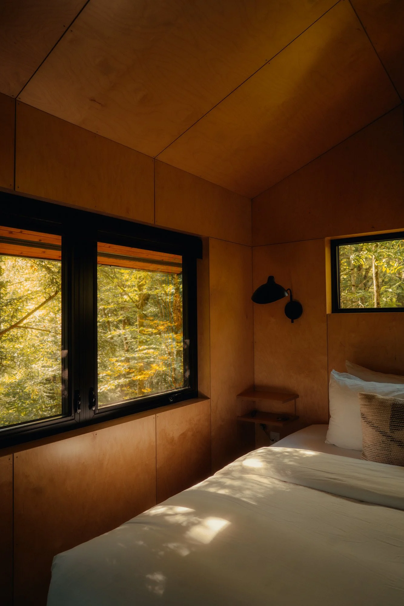 A cozy bedroom with wooden walls and ceiling, featuring a bed with white bedding and pillows, a black reading lamp on the wall, a small wooden nightstand, and windows showing green trees outside with sunlight filtering through the leaves.