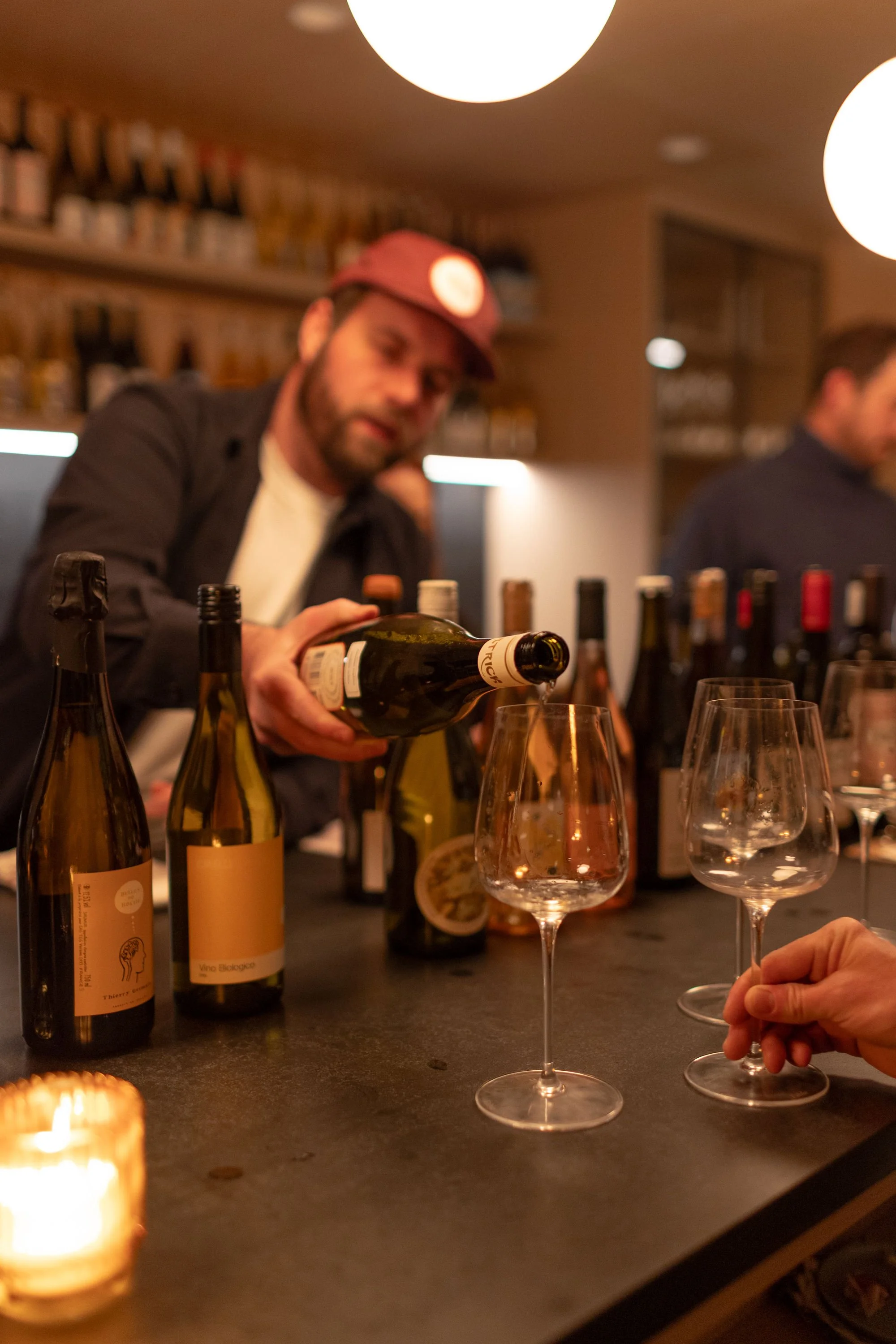 A man pouring wine into a wine glass at a bar with multiple wine bottles and glasses on the counter, and a candle in the foreground.
