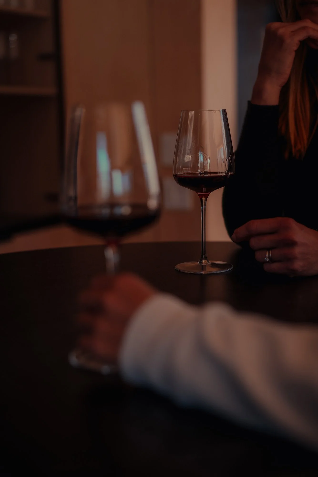 Two glasses of red wine on a dark table, with a woman in a black top holding one glass and a person in a white shirt in the foreground holding a third glass.
