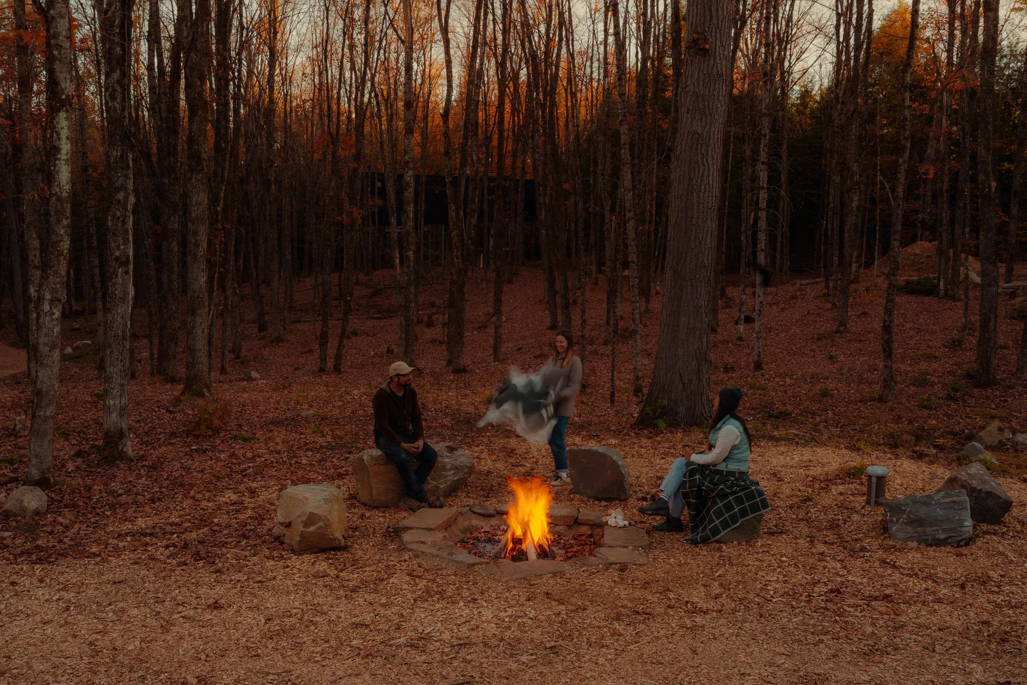 People sitting around a campfire in a wooded area during sunset.