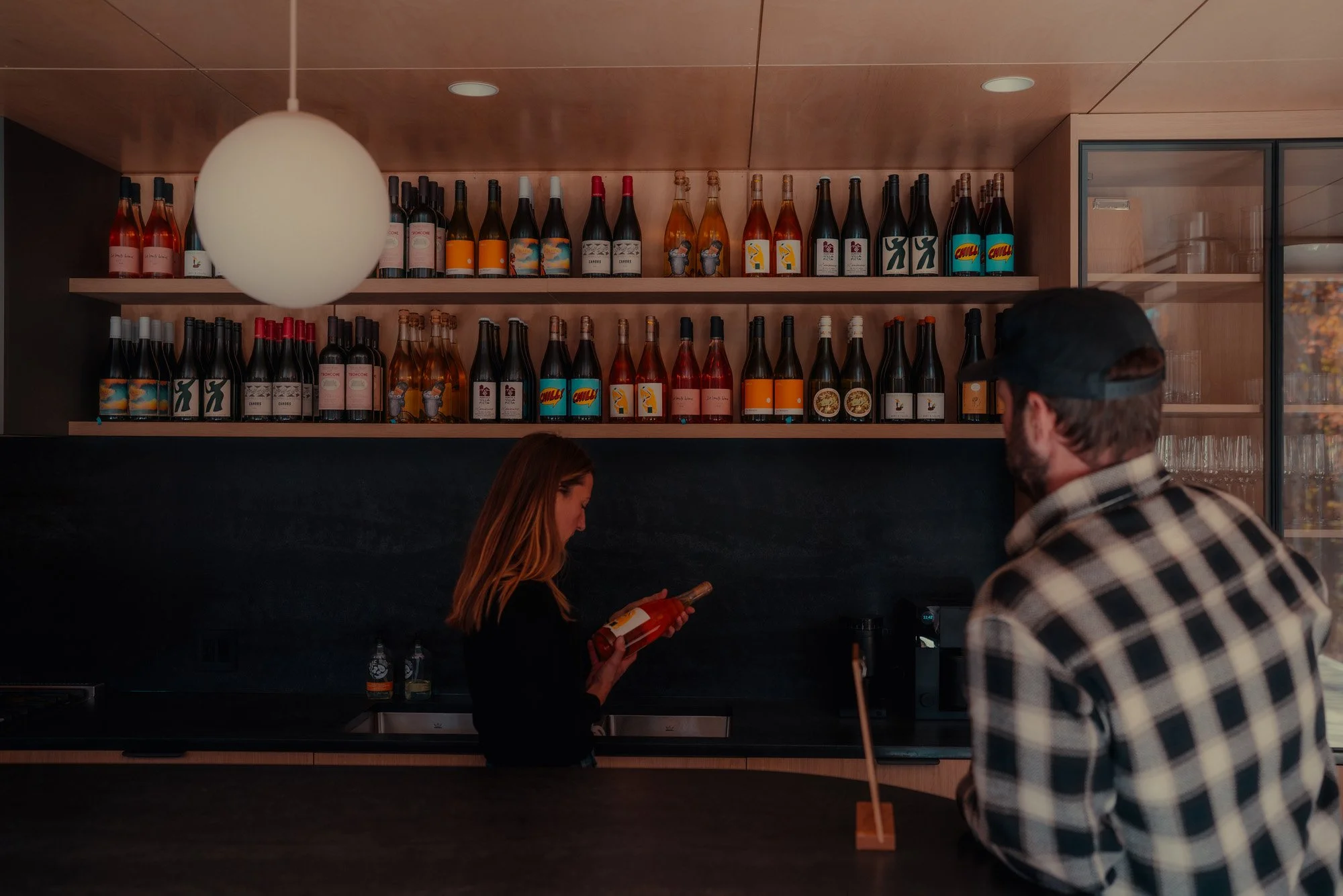 A woman and a man standing at a bar, with shelves of wine bottles behind them and a large hanging light above.