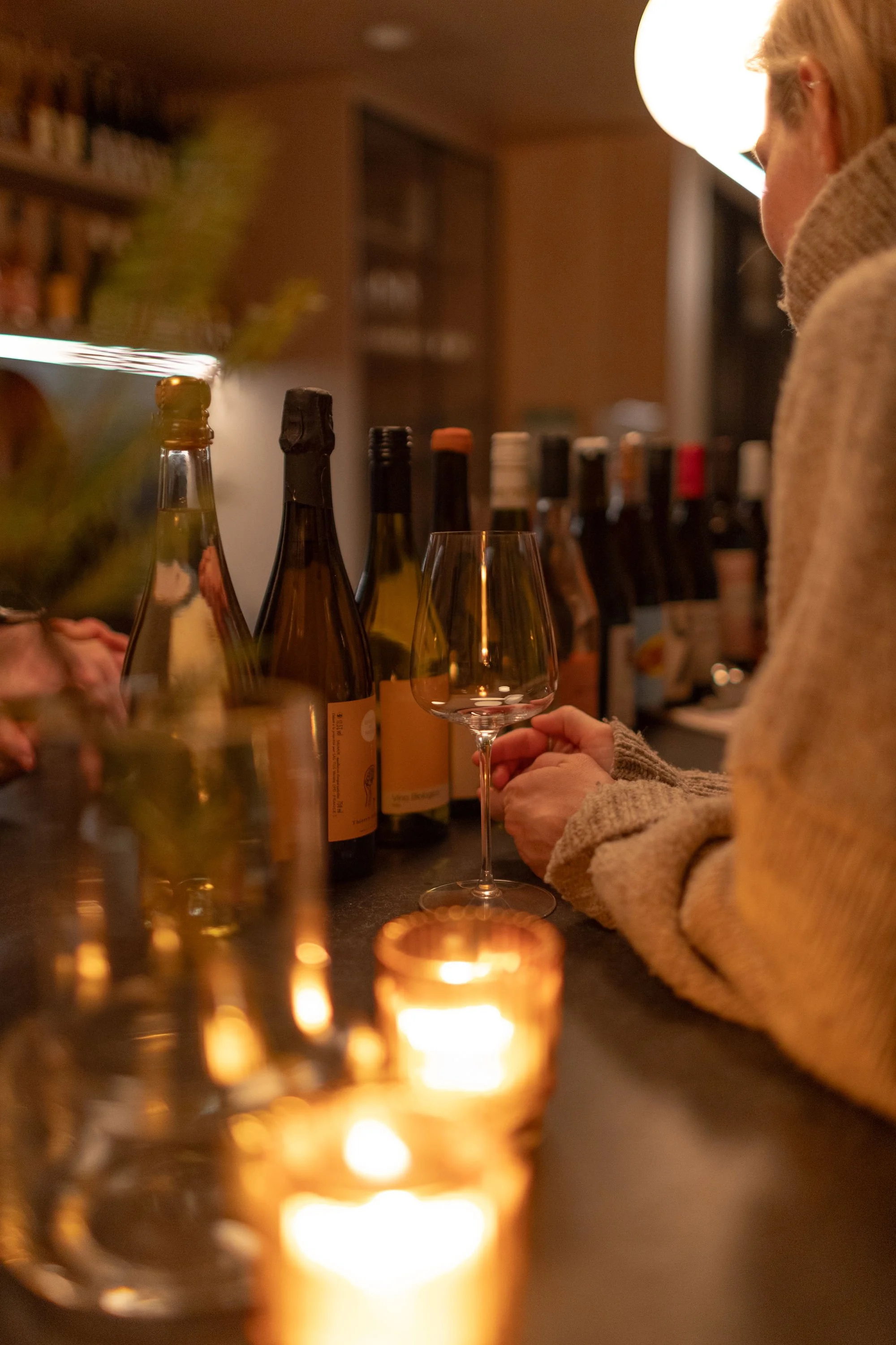 A person in a cozy sweater holding a wine glass at a dimly lit bar or kitchen counter with various bottles of alcohol and a candle.