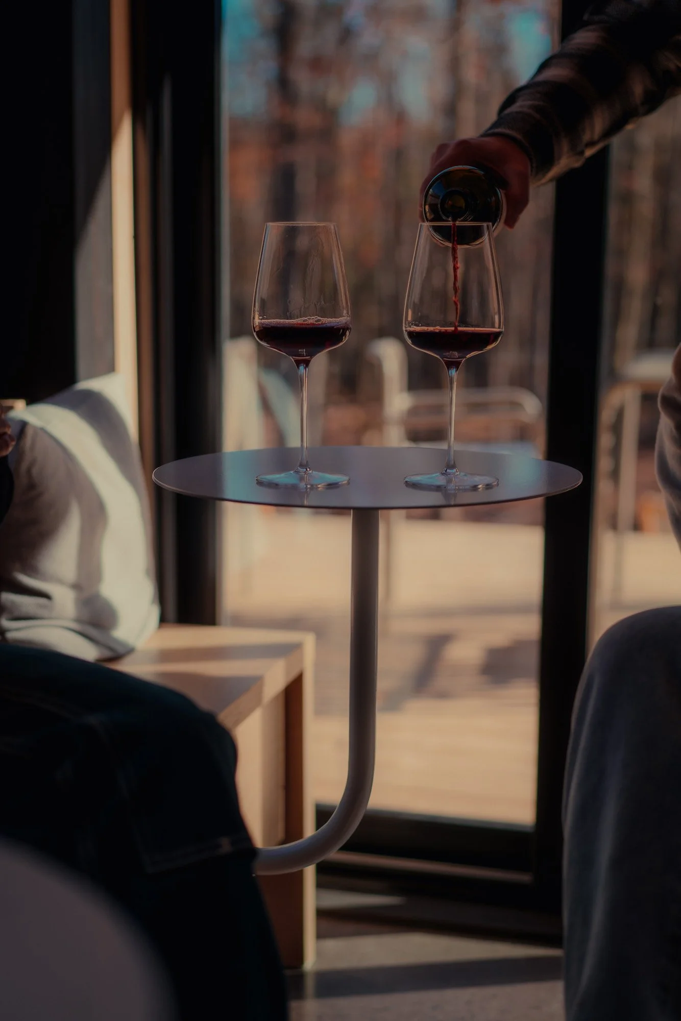 A person pouring red wine from a bottle into a glass, with another filled glass on a small table near a large window, outside trees are visible.