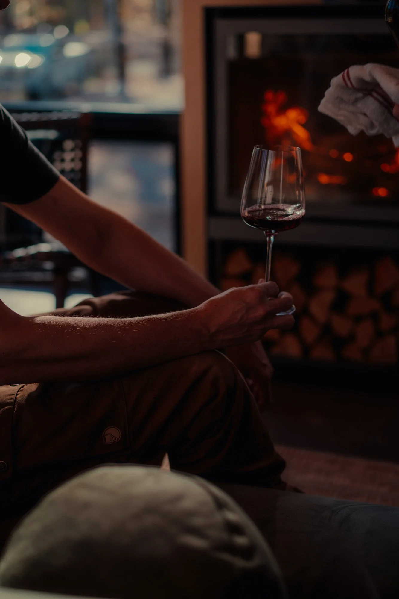 Person holding a glass of red wine near a fireplace in a cozy indoor setting.