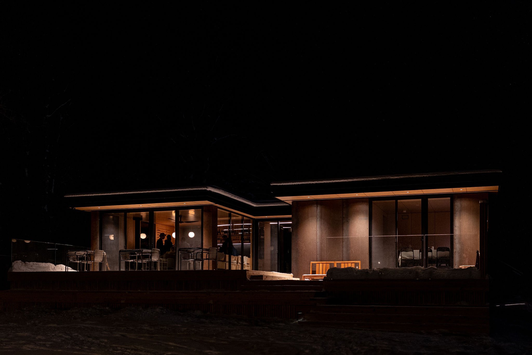 A modern house with large glass windows at night, with warm interior lighting. The house is elevated with steps leading up to it, and appears to have a deck or porch area.