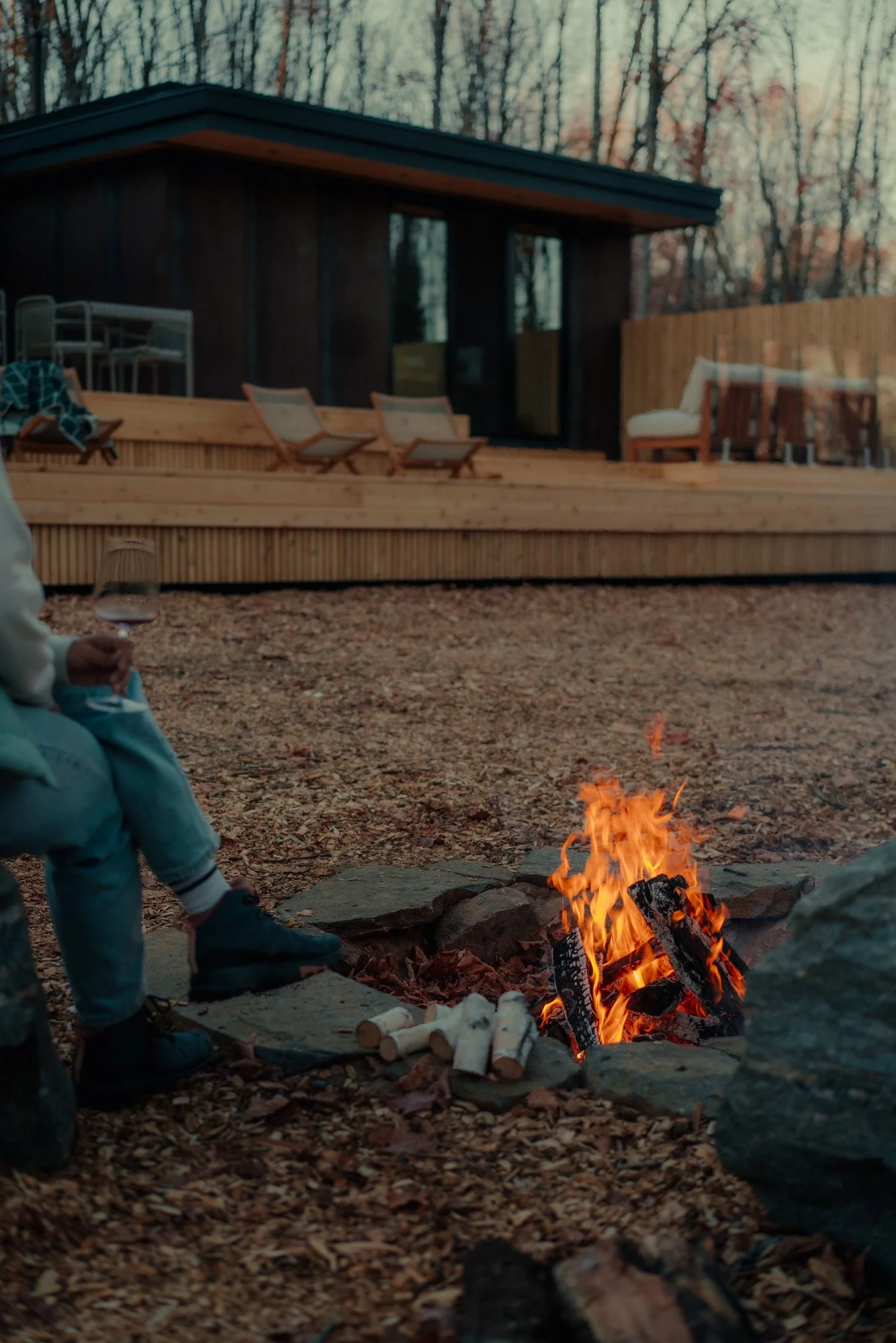 Person sitting by a campfire holding a glass of wine with a modern outdoor deck and house in the background, set in a wooded area during dusk.
