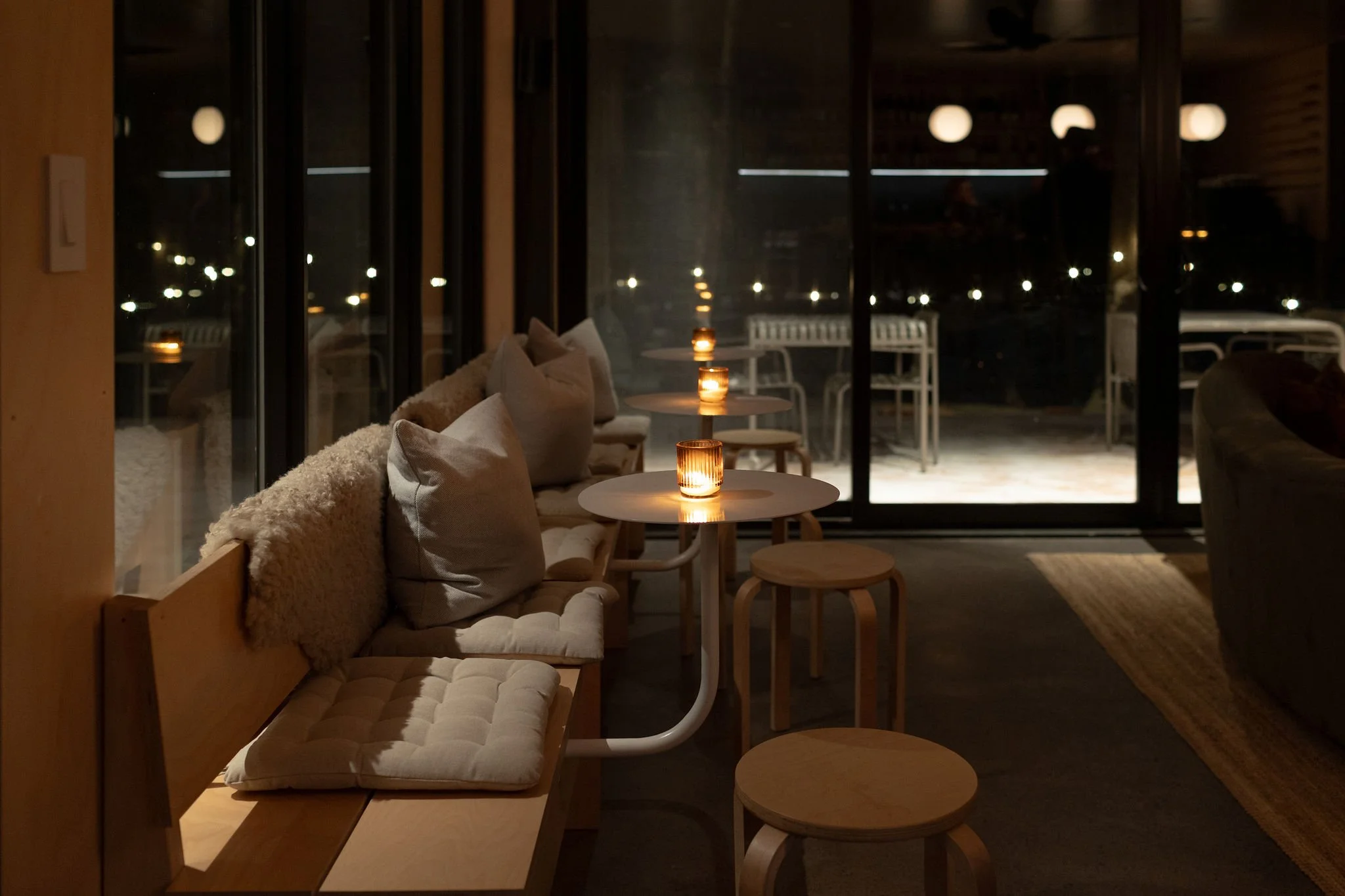 Interior of a cozy cafe with small round tables, candles, a cushioned bench with pillows, and a large window showing an outdoor patio at night.