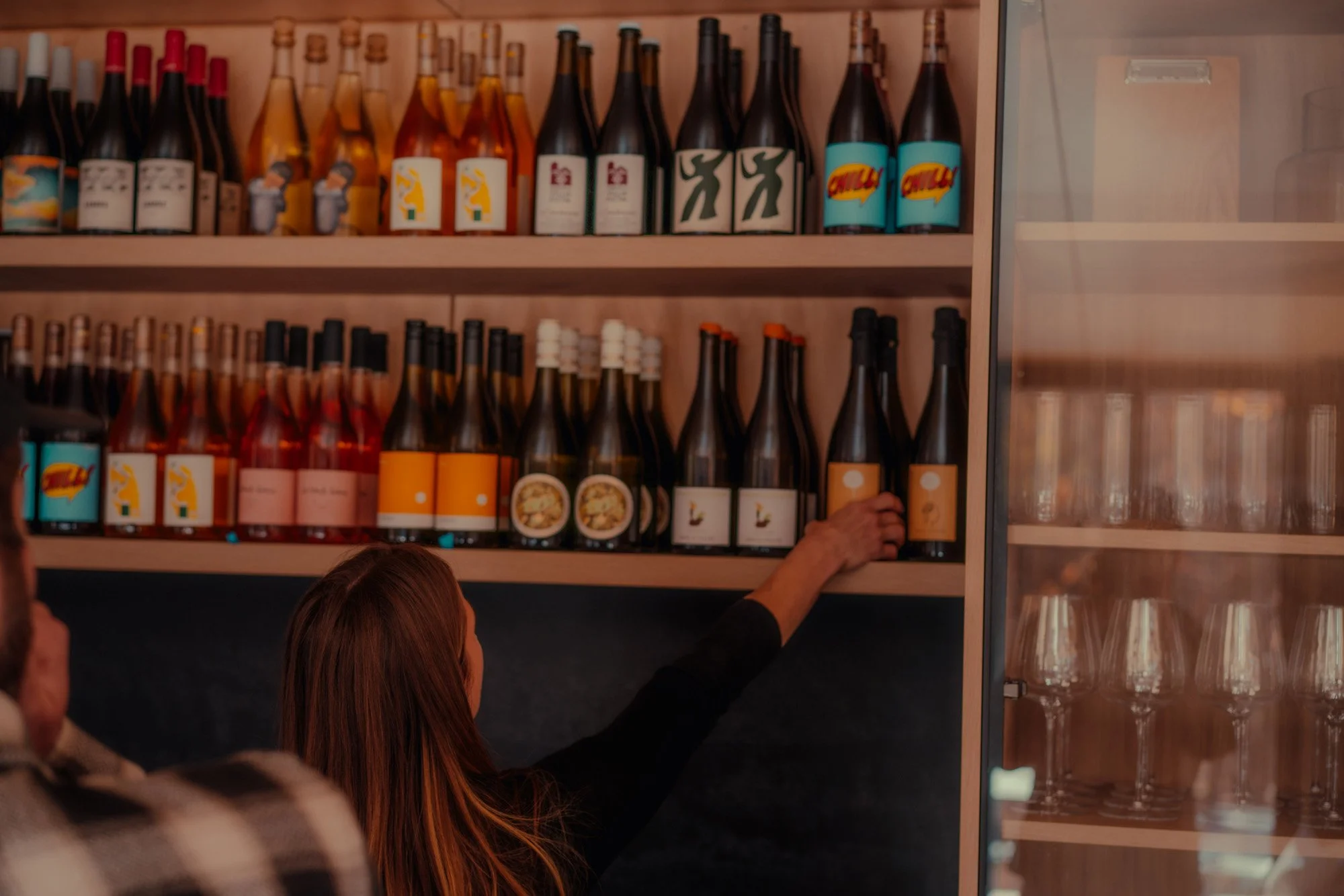 A woman selecting a bottle from a shelf of colorful sauces or condiments in a store or restaurant. The bottles are arranged on wooden shelves with various labels.
