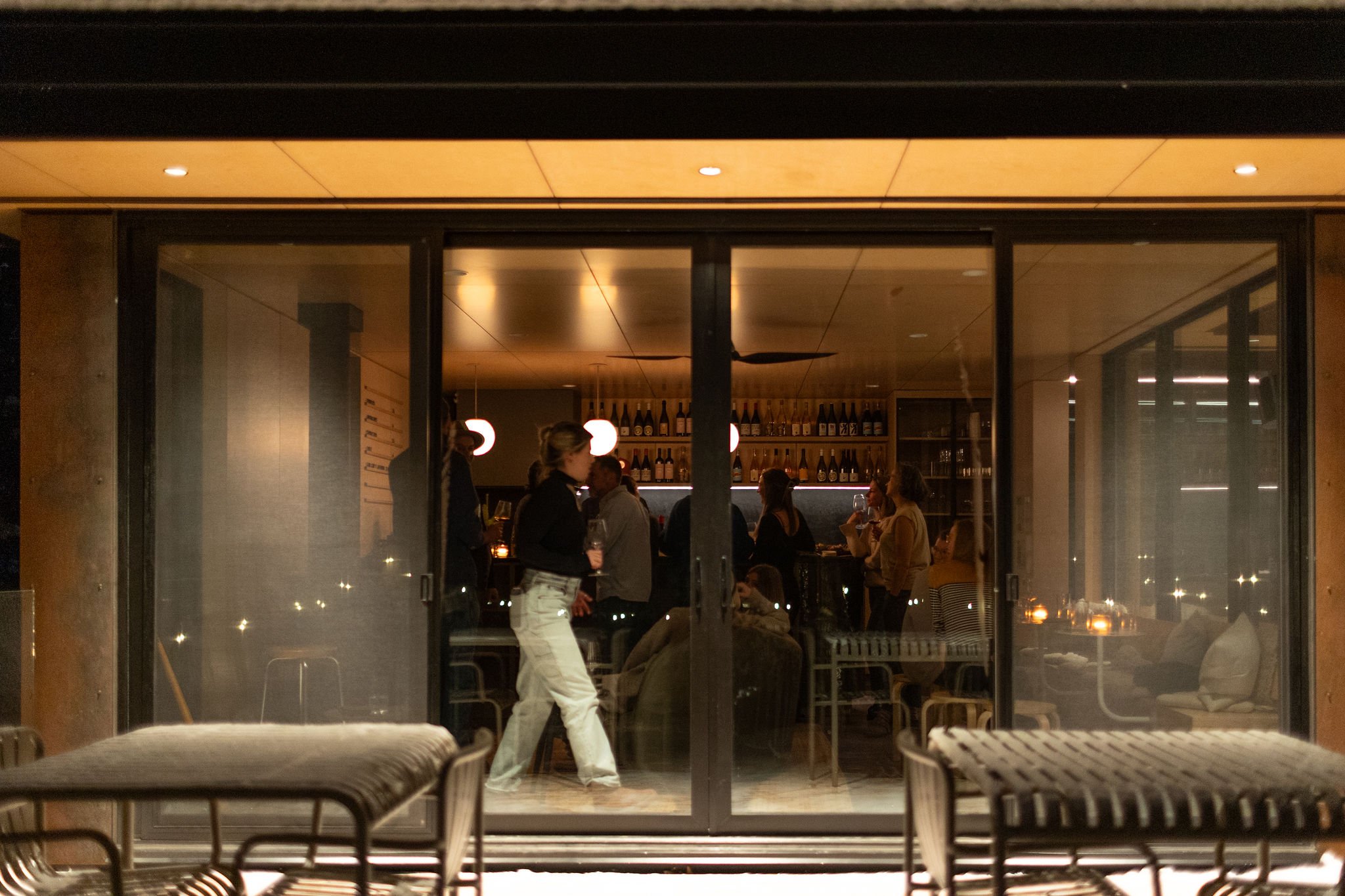 People gather inside a warmly lit bar seen through large glass sliding doors, with outdoor seating in the foreground.