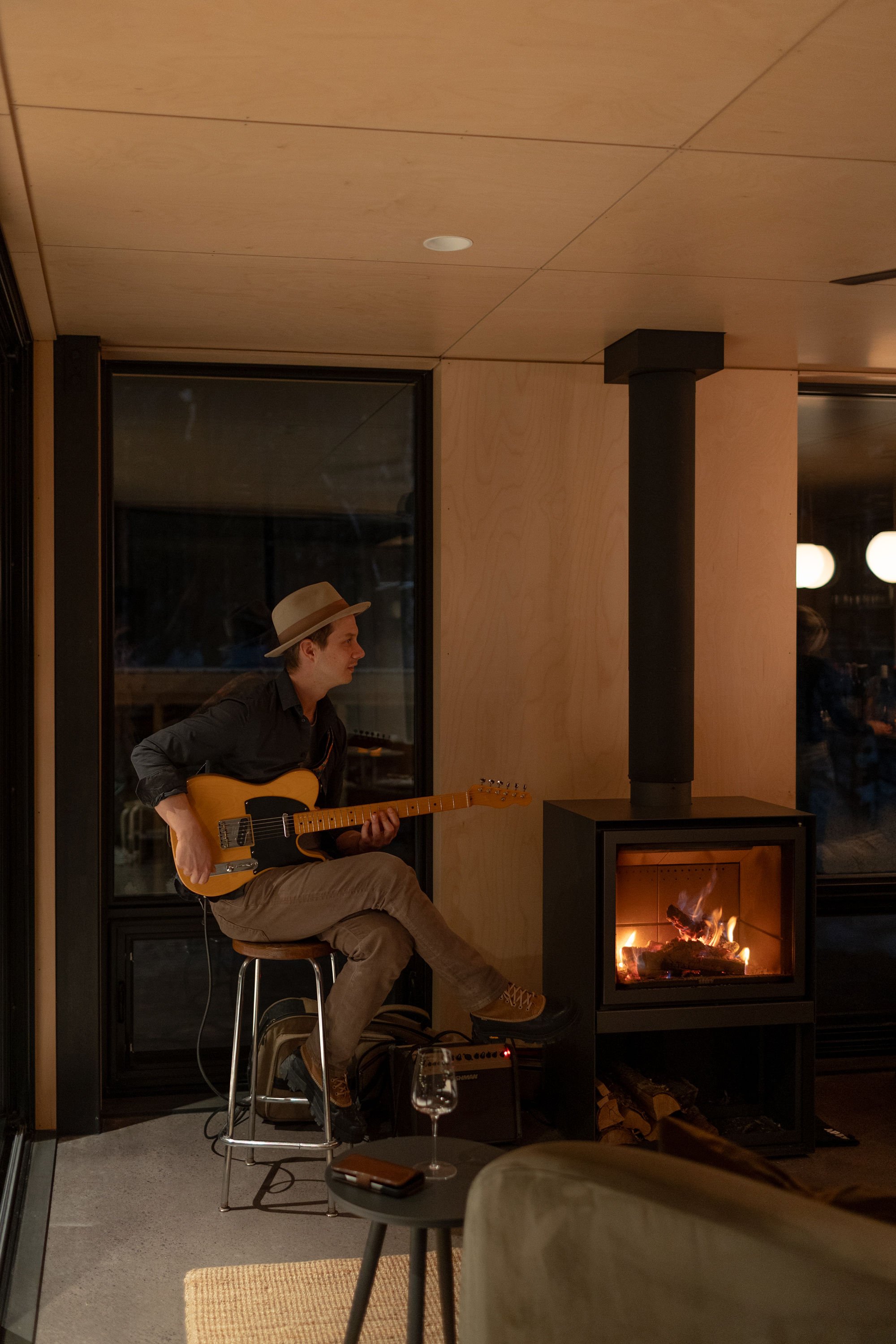 A man wearing a hat and dark shirt plays an electric guitar while seated on a high stool near a fireplace in a cozy indoor setting.