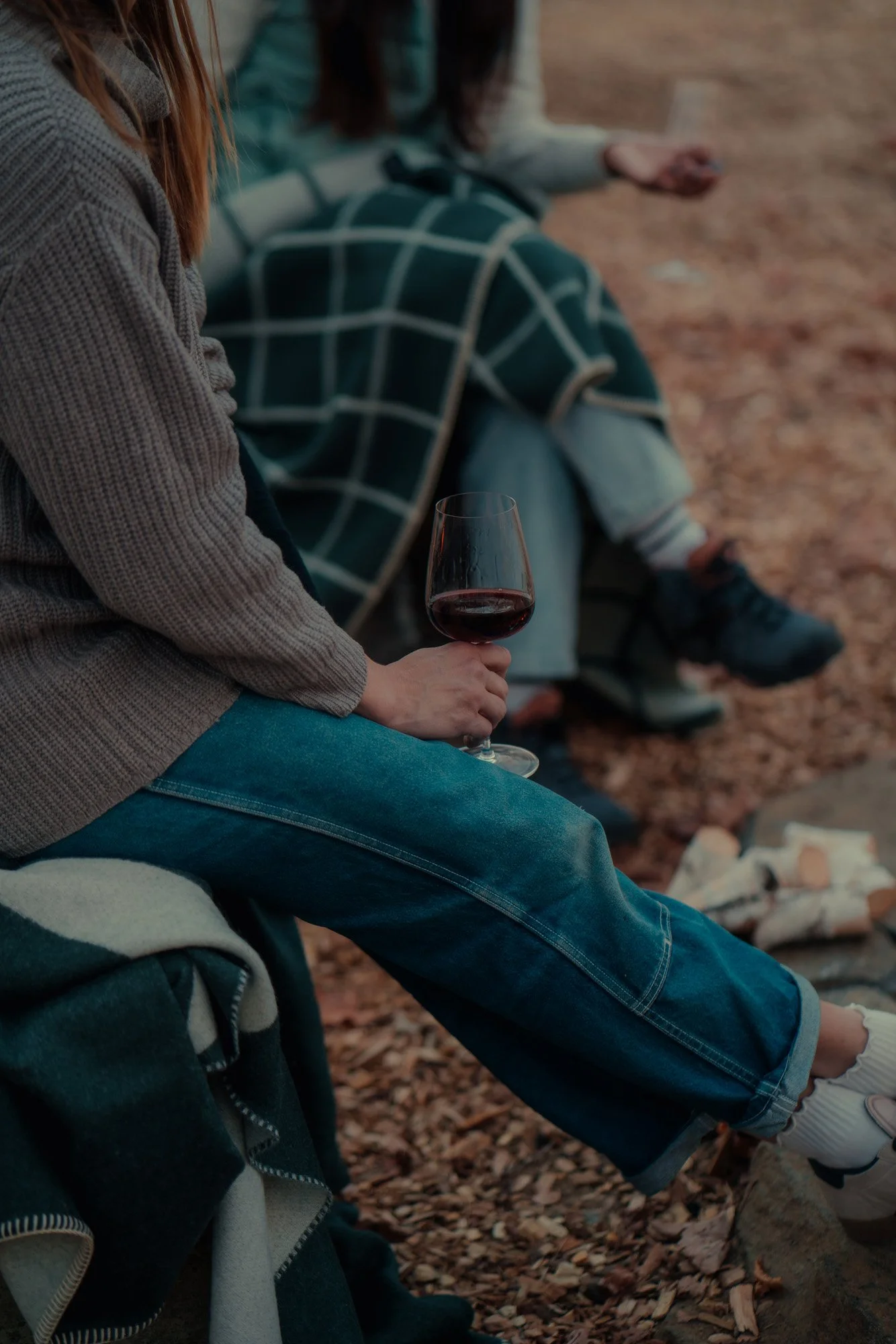 People sitting outdoors on a blanket, holding a glass of red wine, with a campfire in the background.