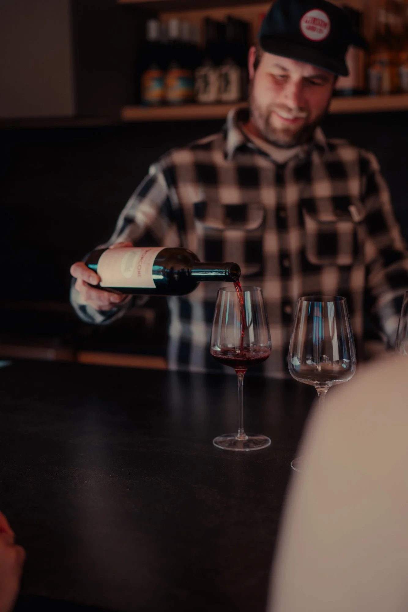 A man wearing a black cap and plaid shirt pouring red wine into a glass at a table, with a second empty wine glass nearby.