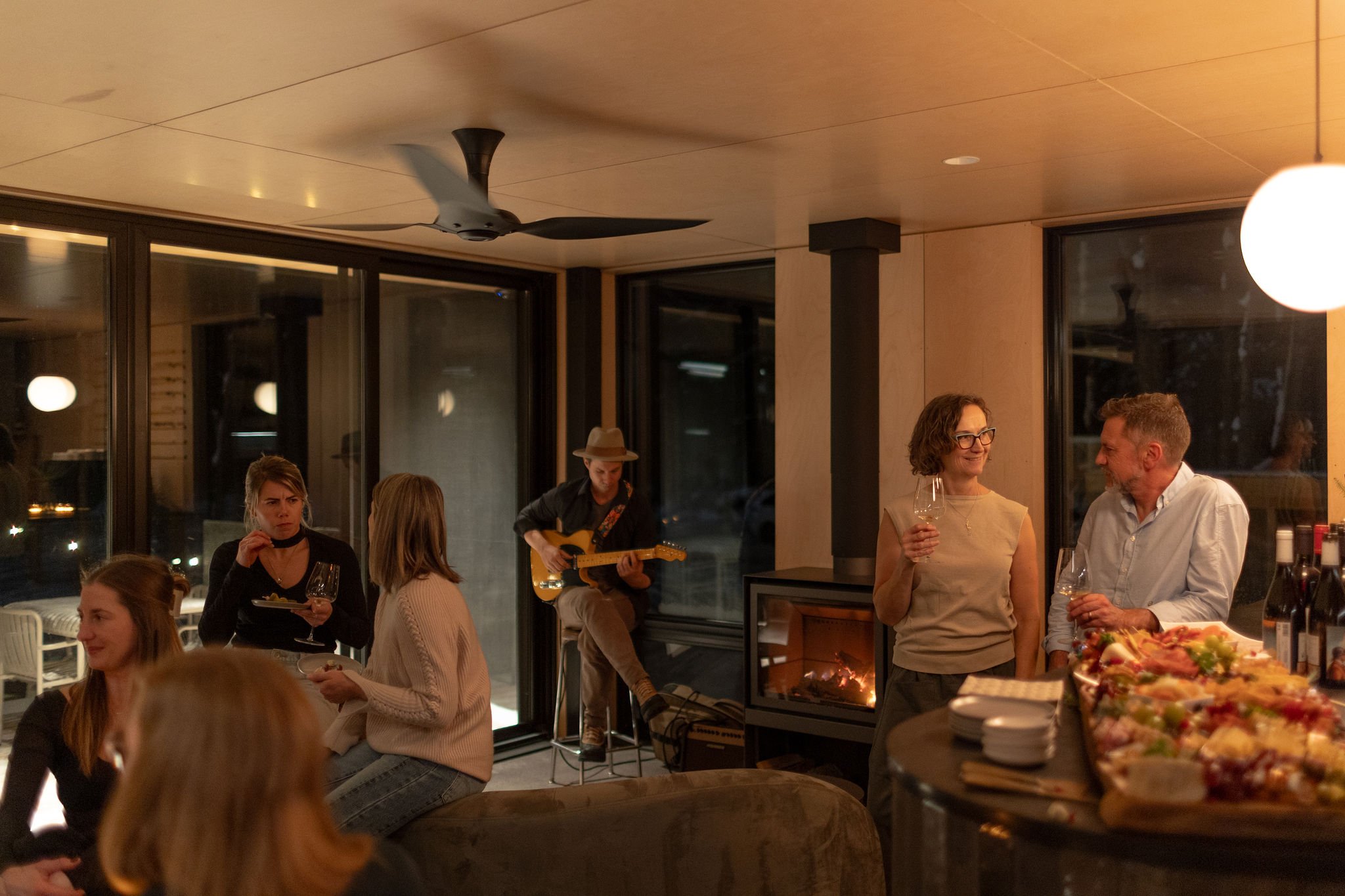 A group of people at a gathering in a cozy indoor space, with some sitting and others standing, holding drinks. A guitarist in a hat plays music near a glass wall, with a lit fireplace adding warmth to the atmosphere.