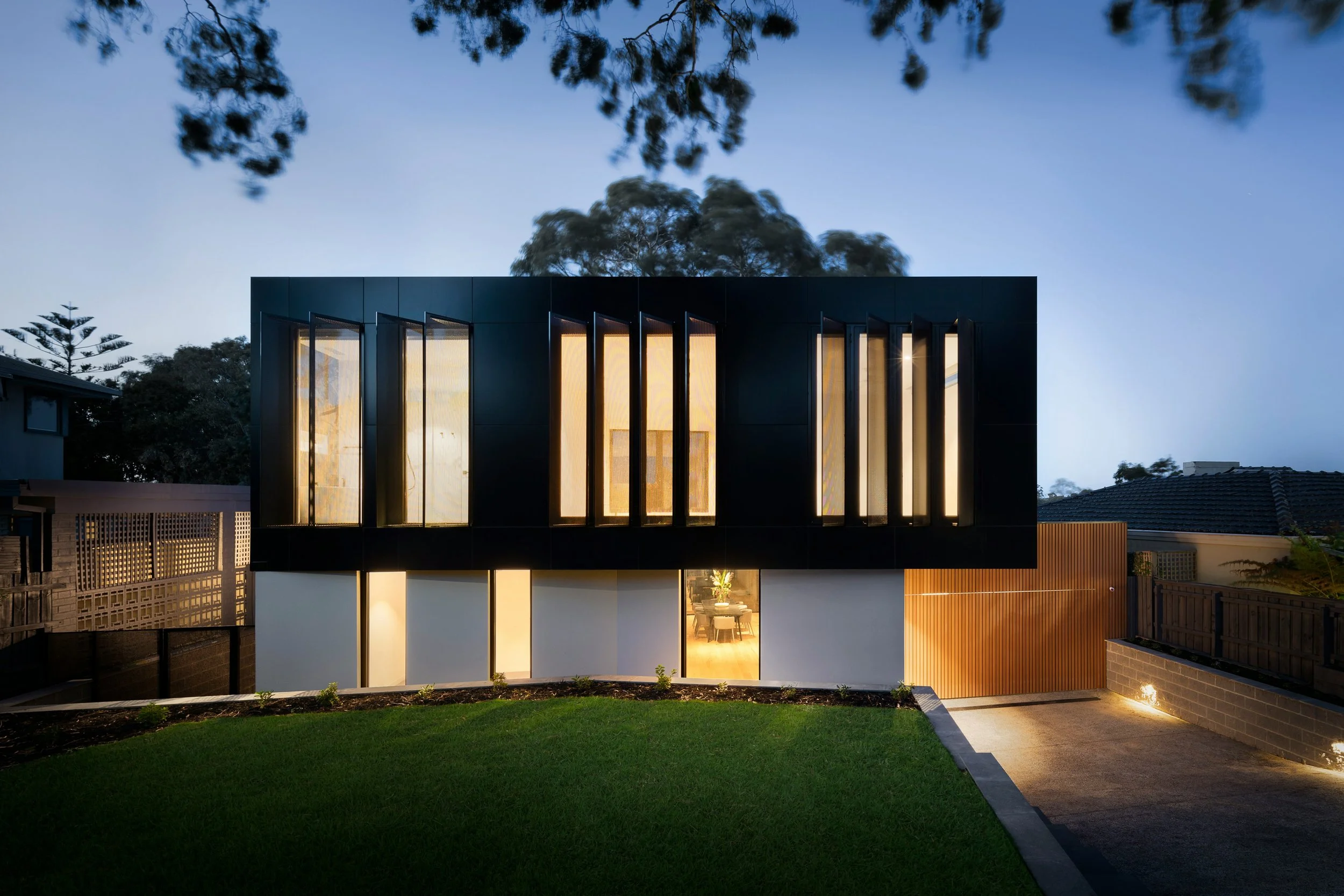 Modern two-story house with black exterior and large vertical windows, illuminated from inside, surrounded by a well-maintained lawn and backyard during early evening.
