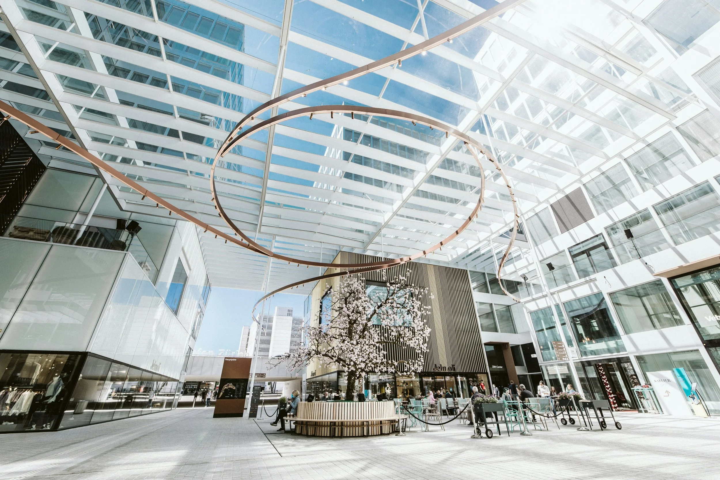 Modern indoor shopping mall with glass ceiling, large spiral copper art installation, and a central planter with blooming cherry blossom tree. People are sitting and walking around, with storefronts and buildings visible through the glass walls.