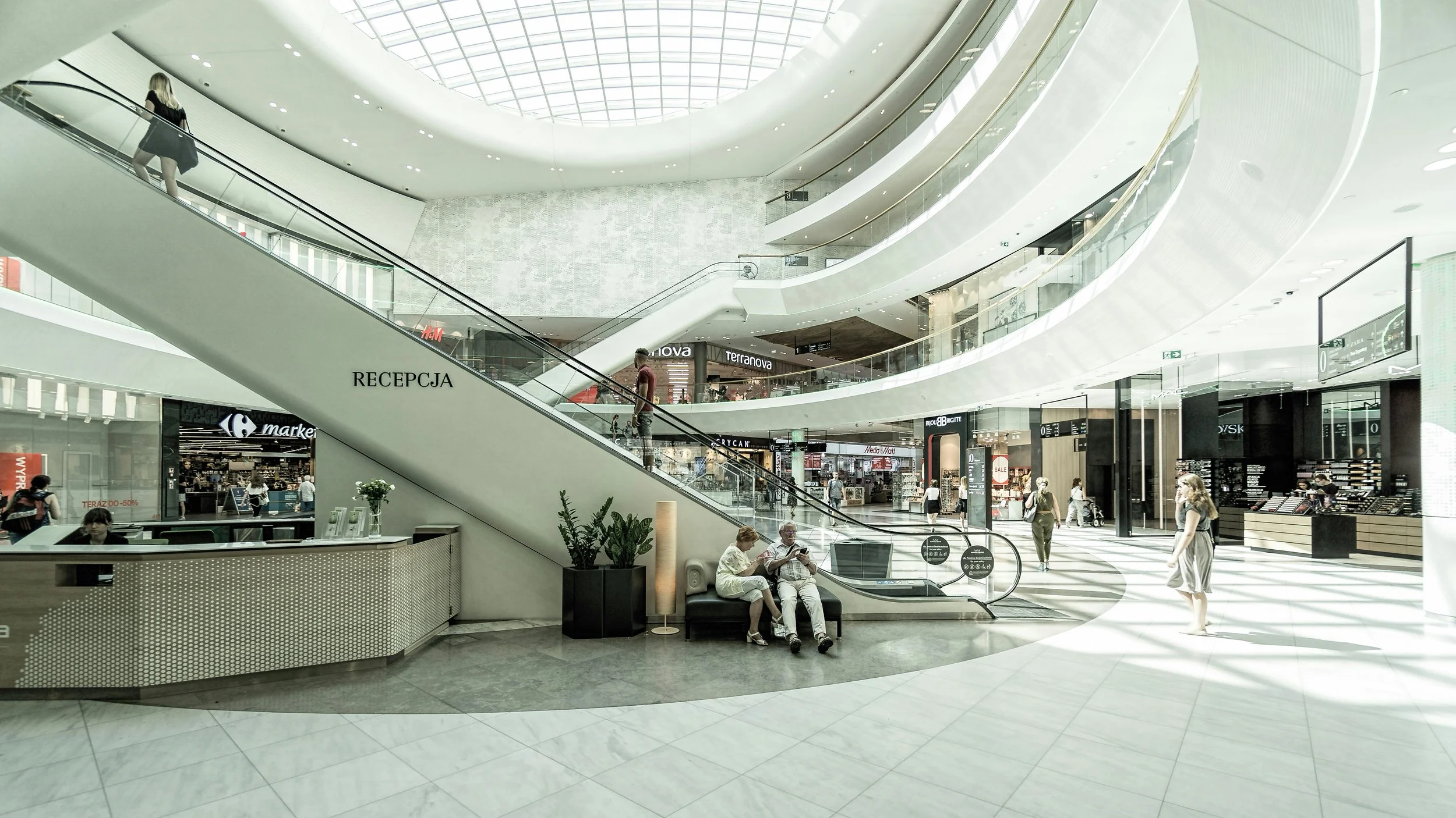 Interior of a modern shopping mall with multiple levels, escalators, shoppers, and storefronts, featuring natural lighting from a large skylight.