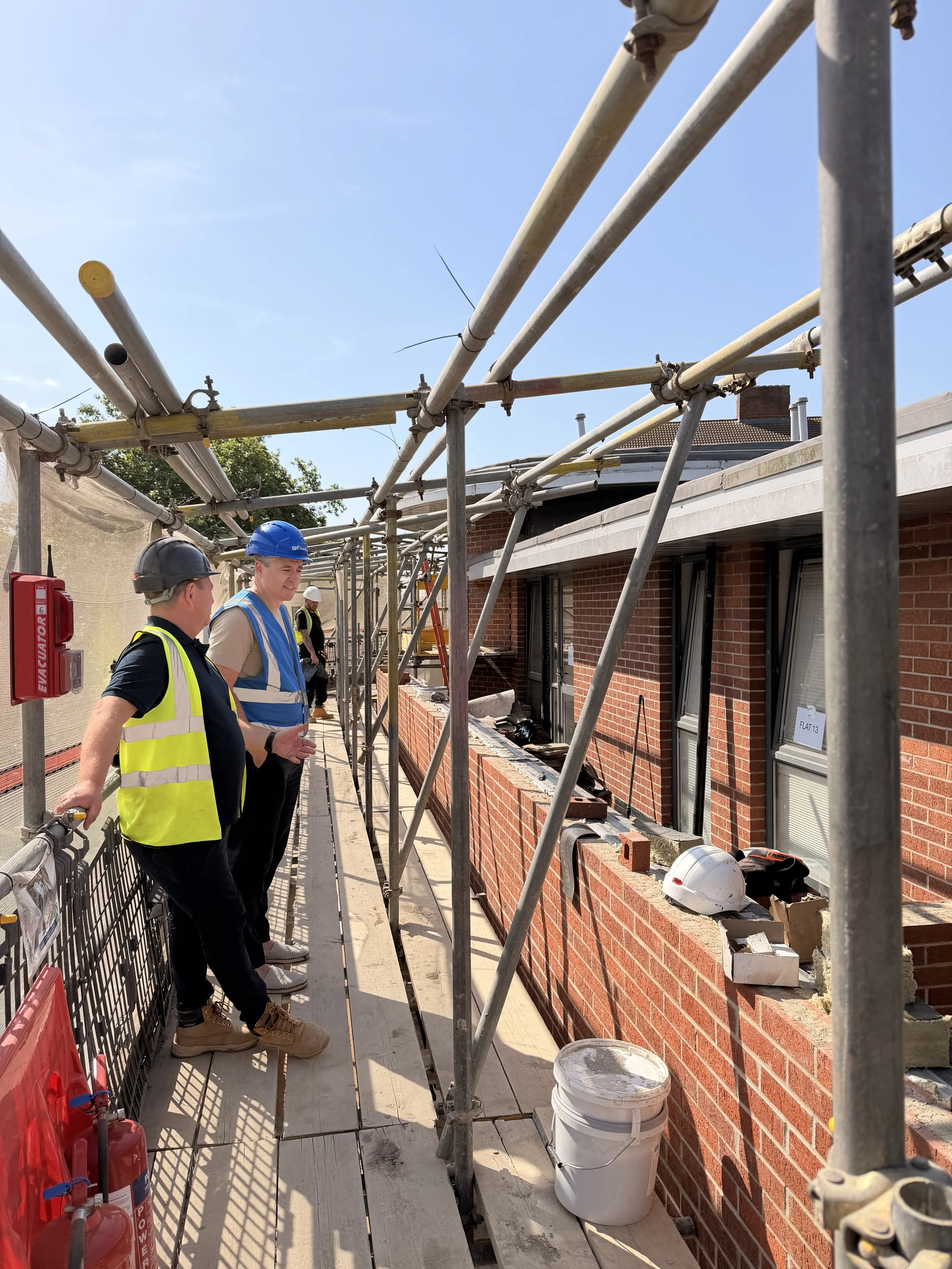 Two construction workers wearing helmets and safety vests stand on scaffolding outside a brick building under construction, with tools and safety equipment nearby.