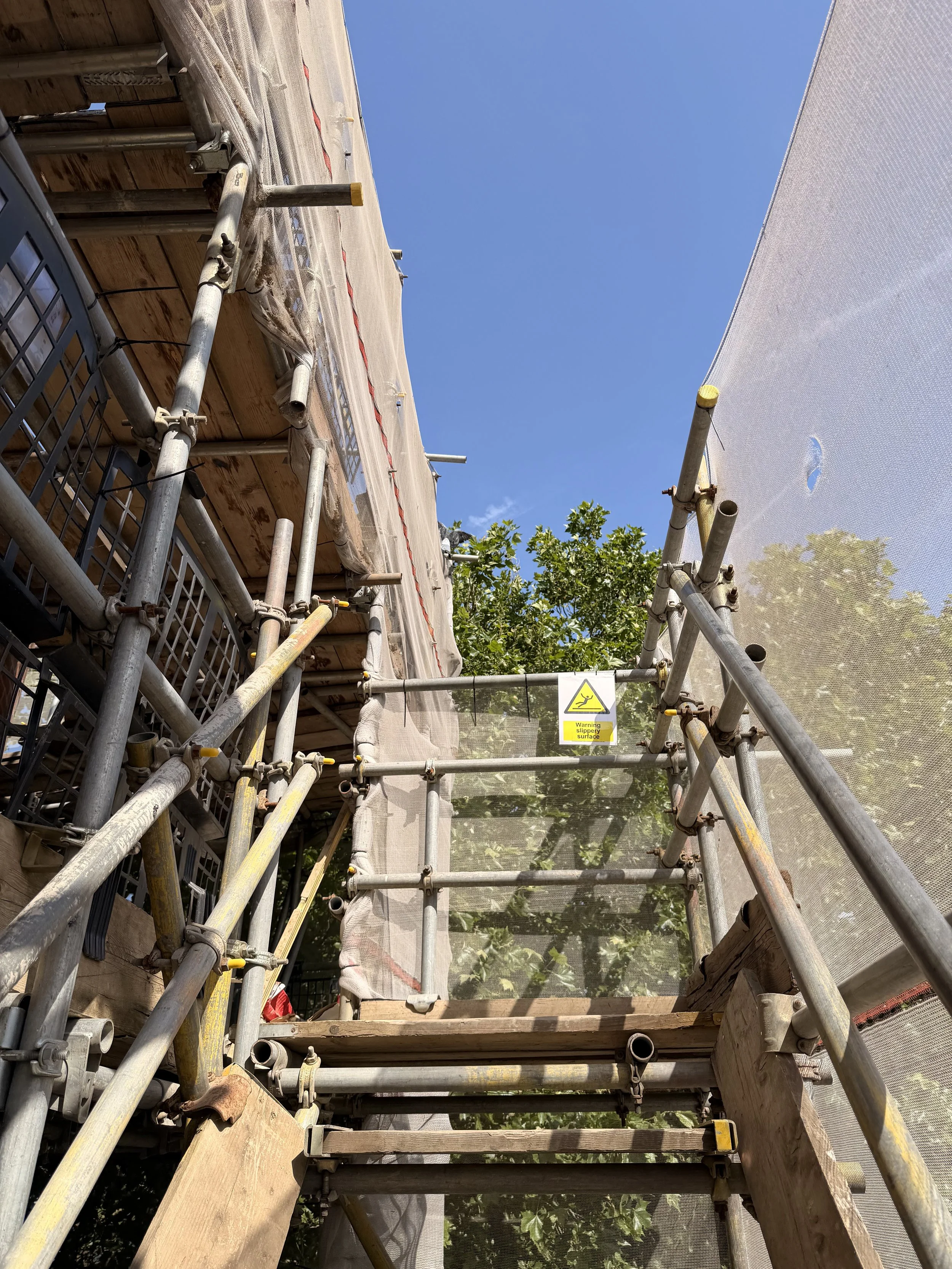 Construction scaffolding with safety netting under a clear blue sky, with some green trees visible in the background.