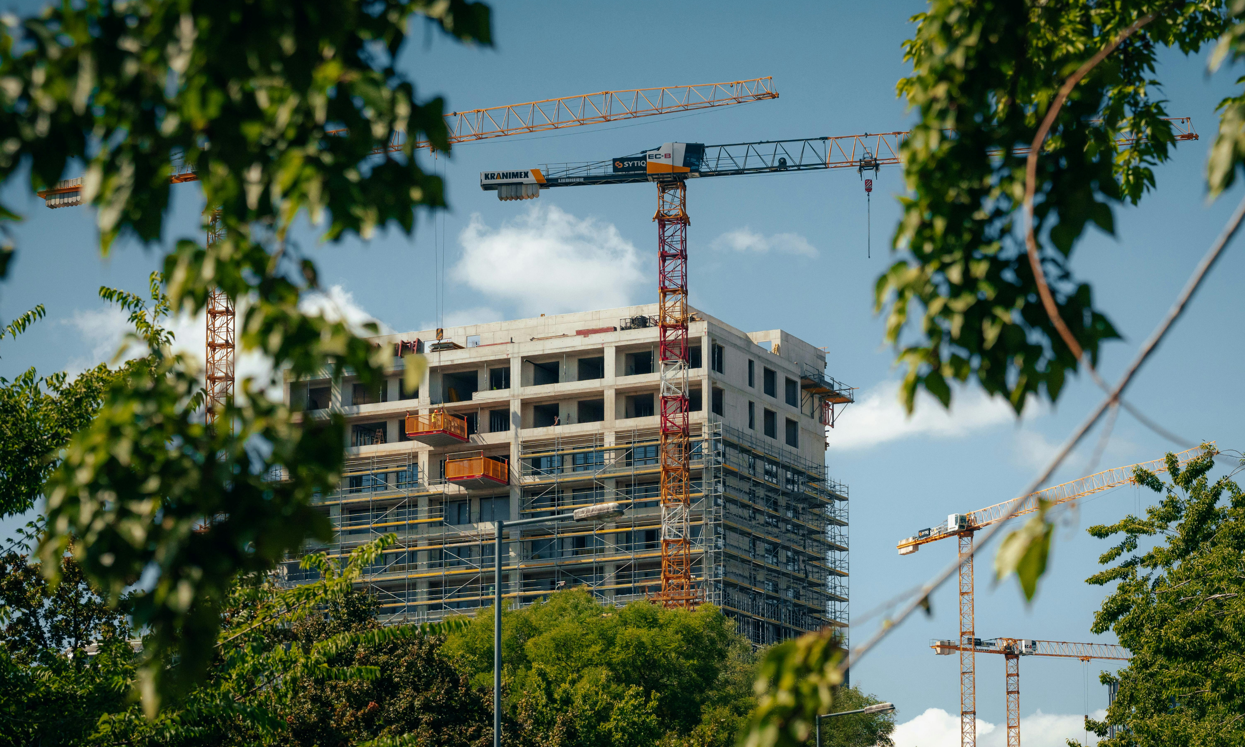 A mid-rise residential building under construction, surrounded by scaffolding and tower cranes against a blue sky, viewed through tree foliage.