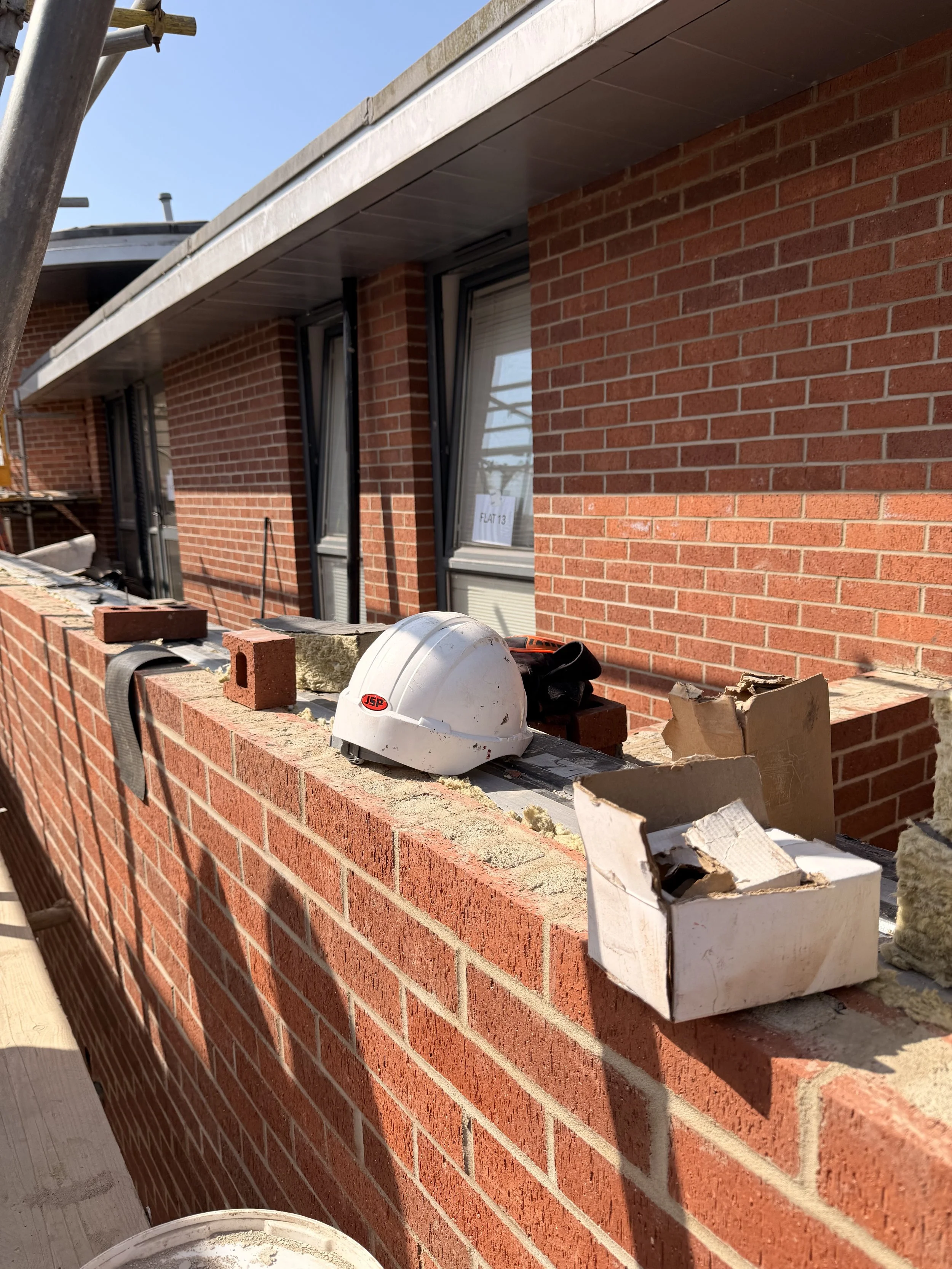 View of a brick building under construction, with scaffolding and building tools on the ledge, including a white hard hat, a white box, and bricks.