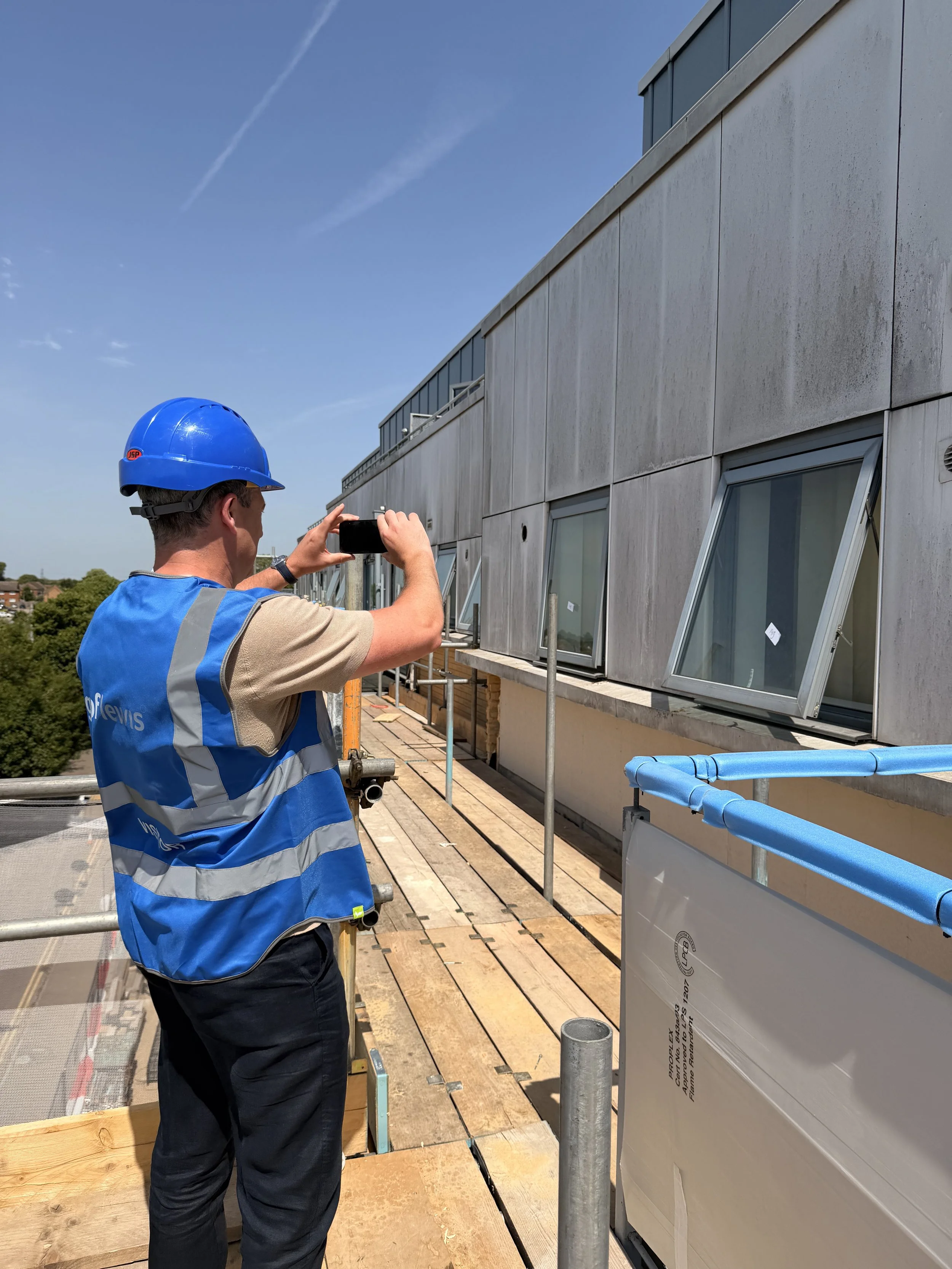 Philip Mallon is wearing a blue safety helmet and a high-visibility vest, taking a photo on a building construction site with windows and scaffolding.