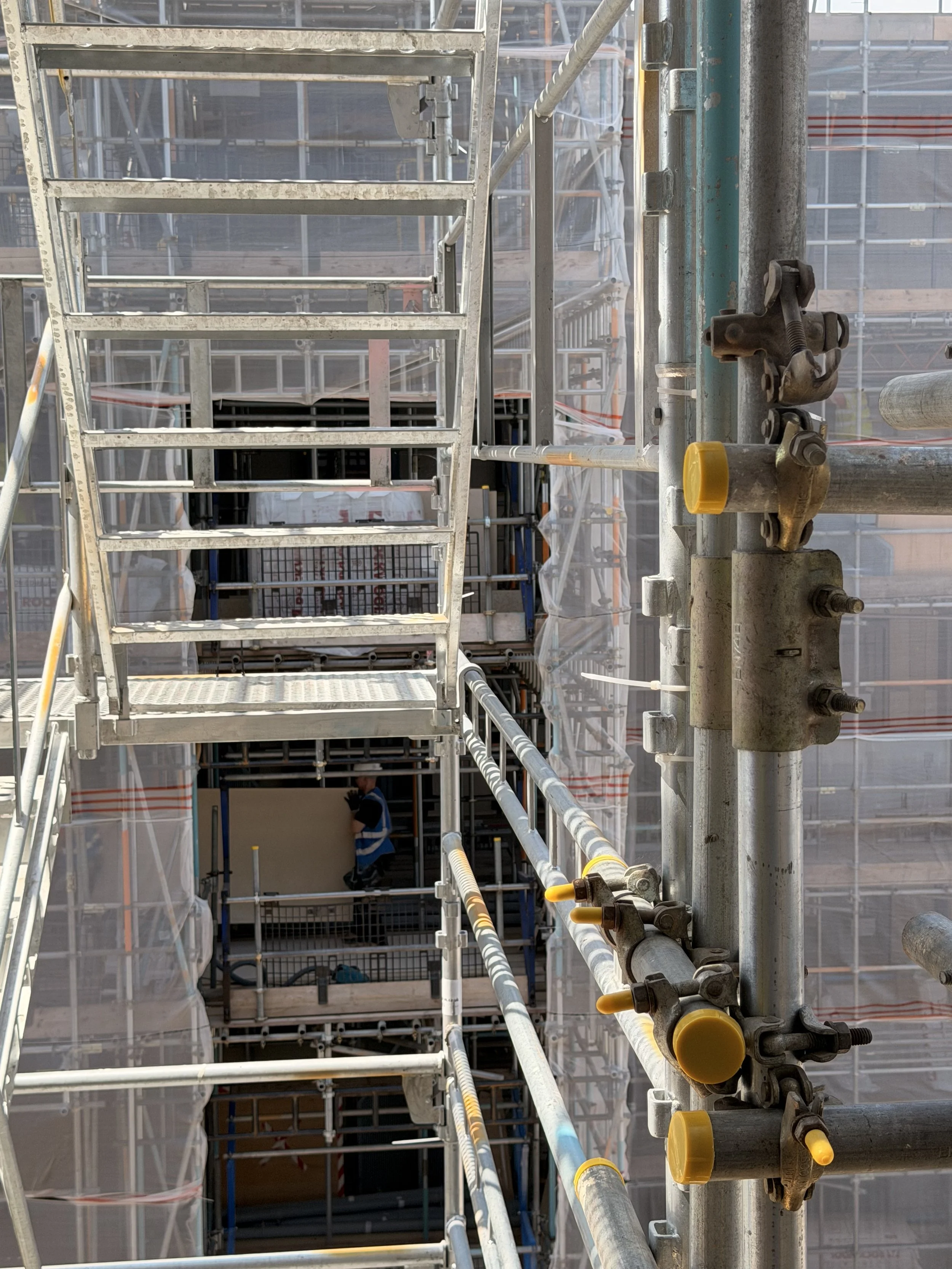 Close-up view of scaffolding at a construction site with metal pipes, clamps, and safety barriers, with partial view of a worker wearing a helmet and safety vest in the background.