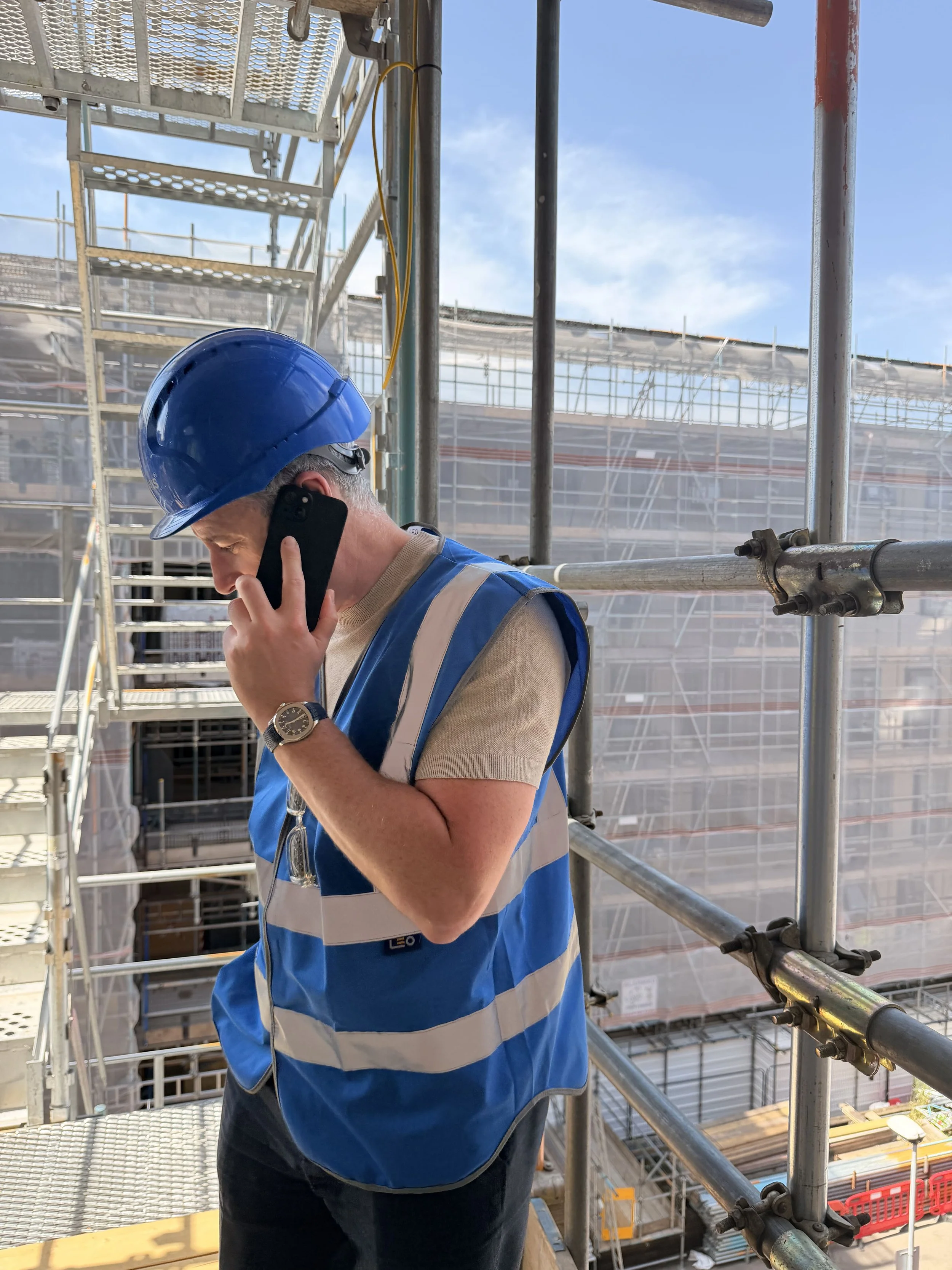 Philip Mallon stands in a blue safety vest and hard hat talking on a mobile phone at a building site with scaffolding and a partly built structure in the background.